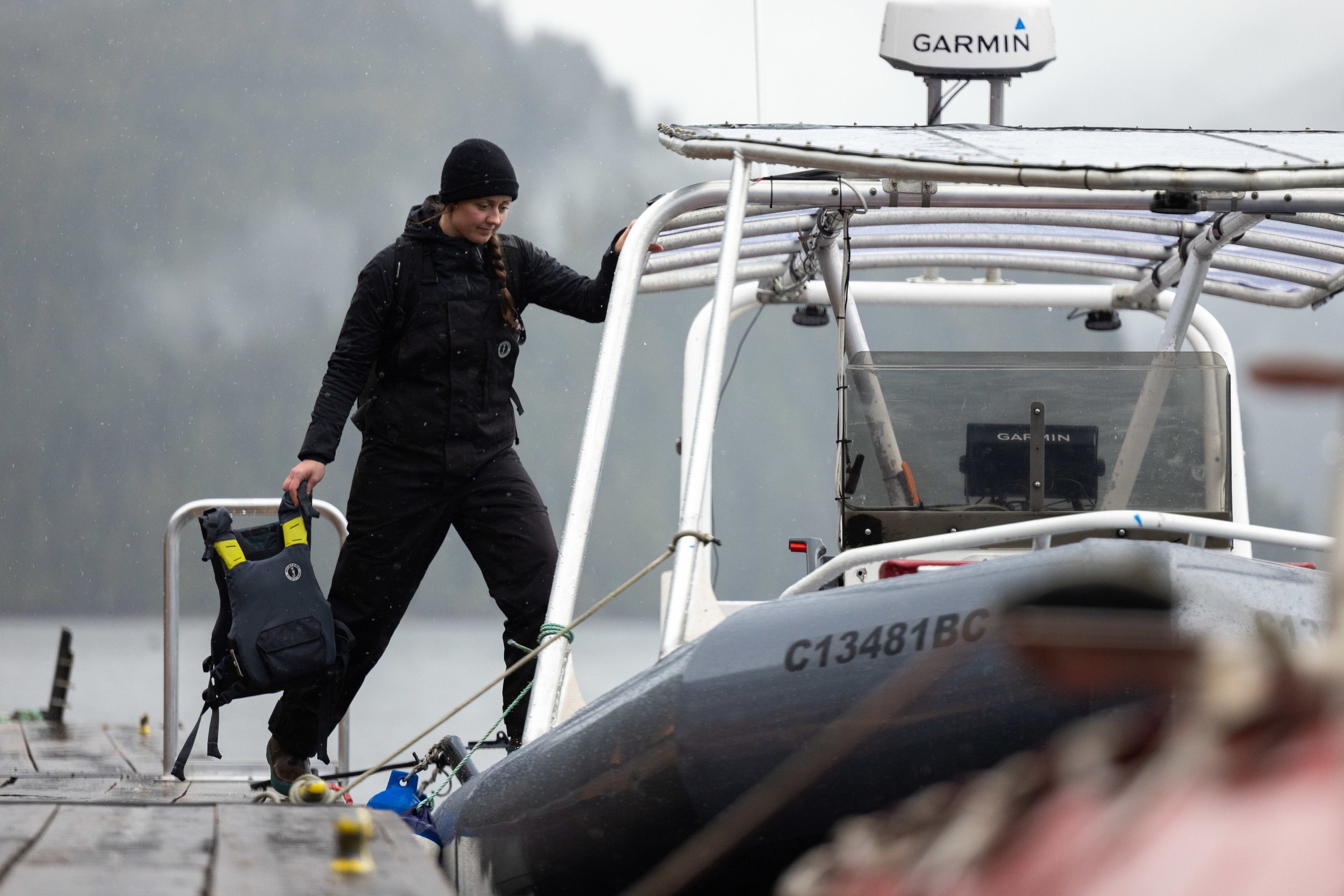 A woman in black rain gear holds a PFD while stepping from a wooden dock onto an inflatable boat in misty weather.