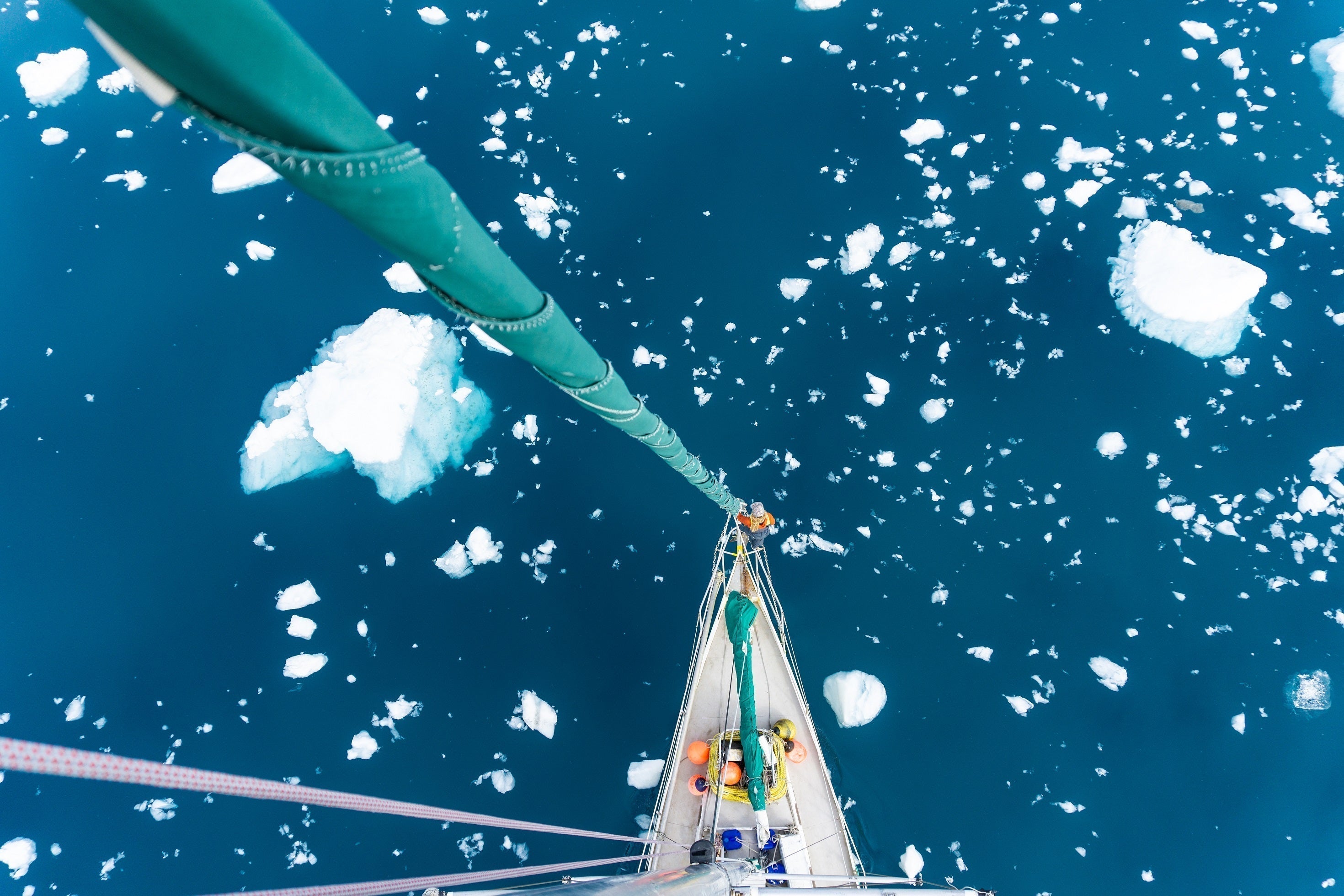 A high-angle, top-down view looking down a sailboat's mast toward the bow as it navigates through deep blue water filled with scattered chunks of sea ice.
