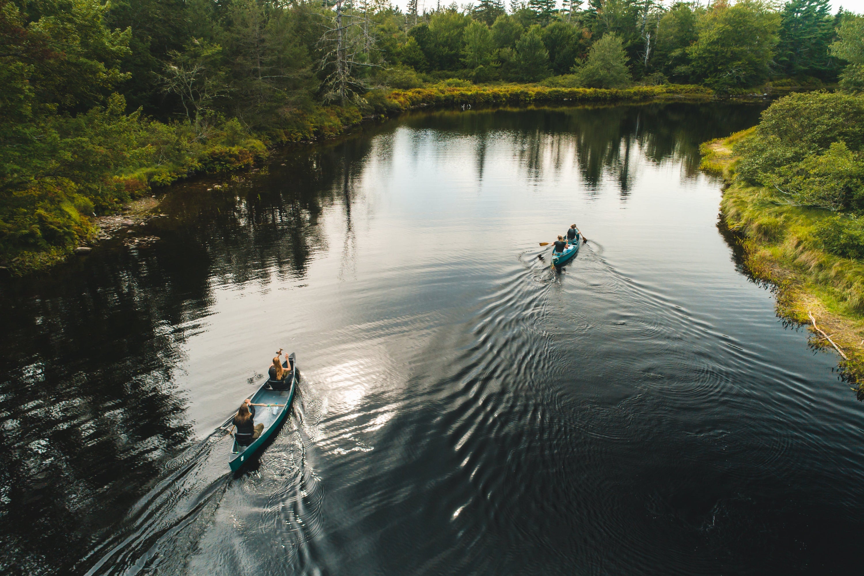 A high-angle aerial view shows two canoes paddling down a calm, reflective river flanked by dense green forests.