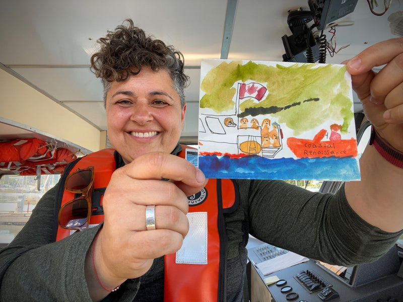 A smiling woman, Debora DeNapoli, wears an orange life vest and holds up a child's watercolor drawing depicting a rescue boat scene.