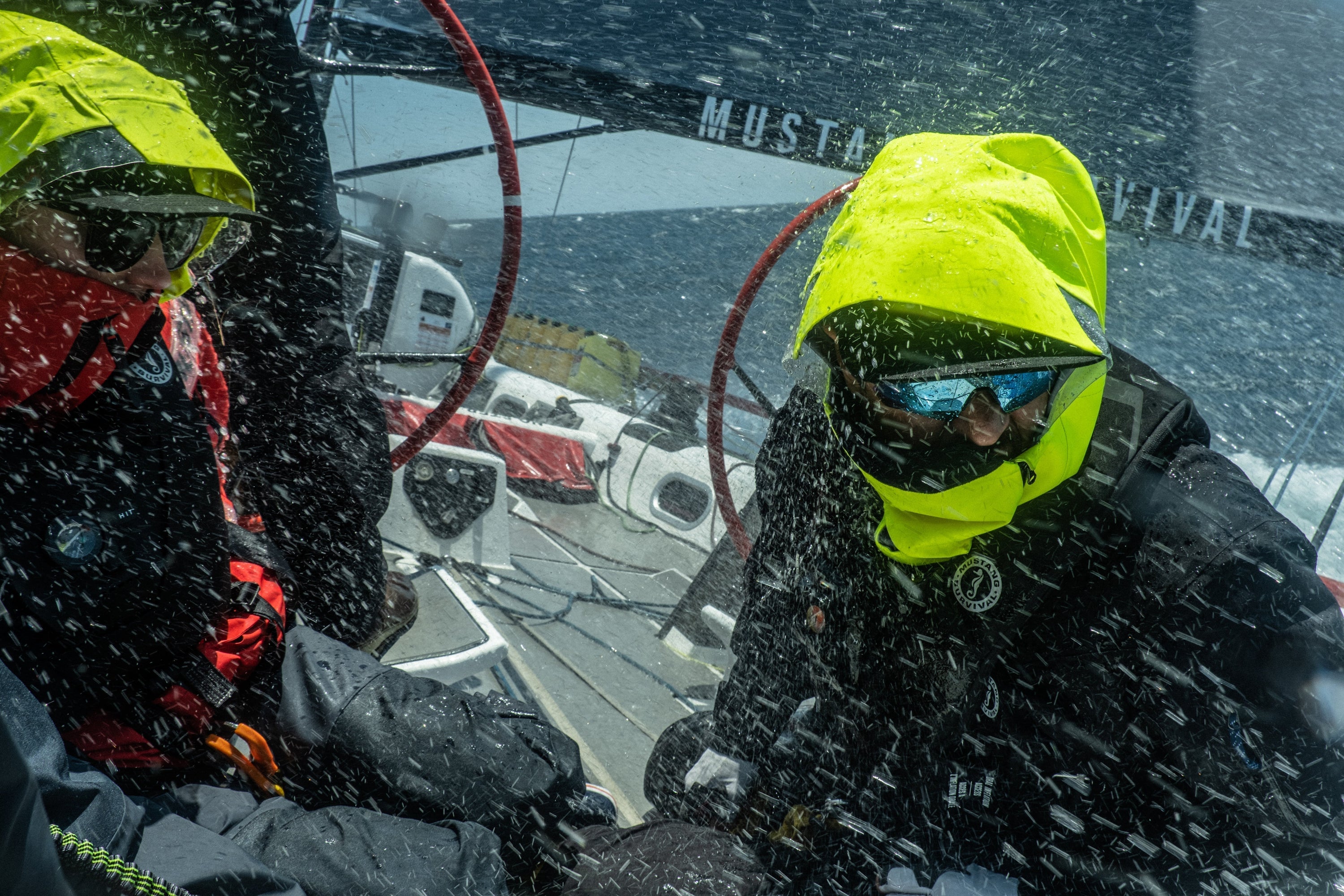 Two sailors on a racing yacht endure heavy ocean spray while wearing high-visibility foul weather gear and hoods.