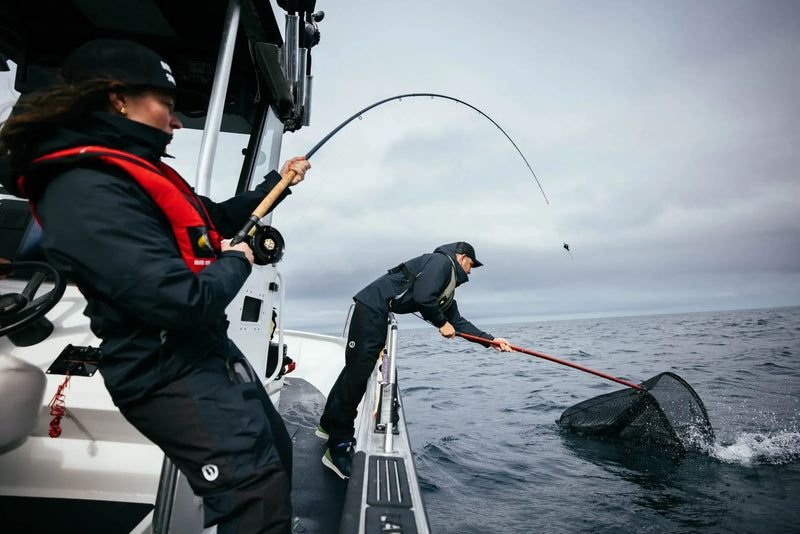 Anglers working together on a boat as one reels in a fish and the other prepares a landing net.