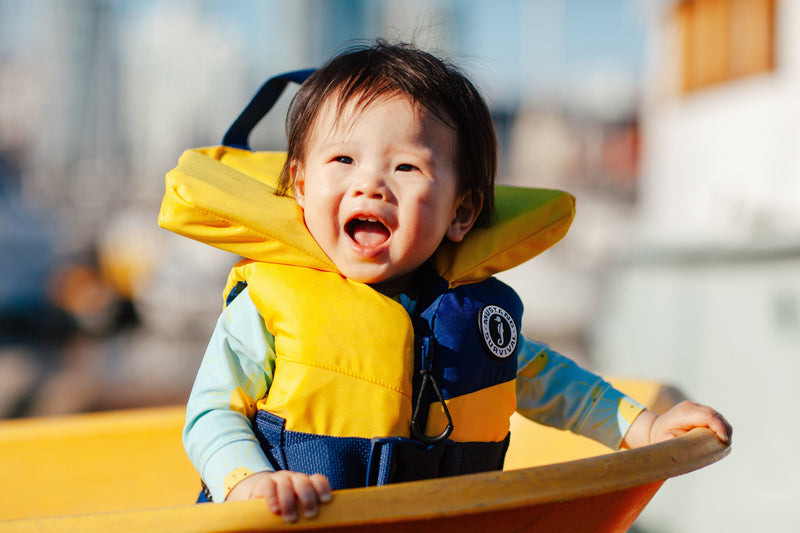A cheerful infant wearing a yellow and blue Lil' Legends PFD smiles while sitting inside a small boat.
