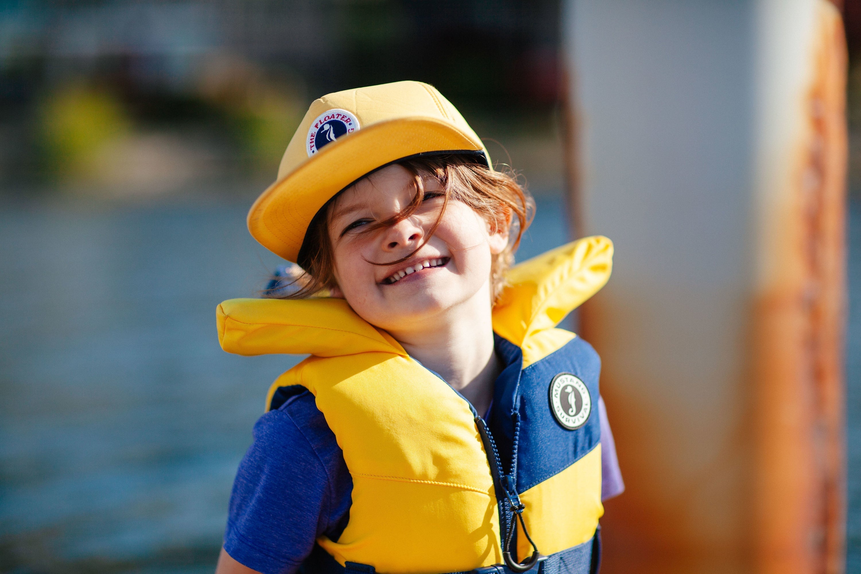 A smiling child on a dock wearing a yellow and blue Lil Legends PFD with a head pillow and safety strap.