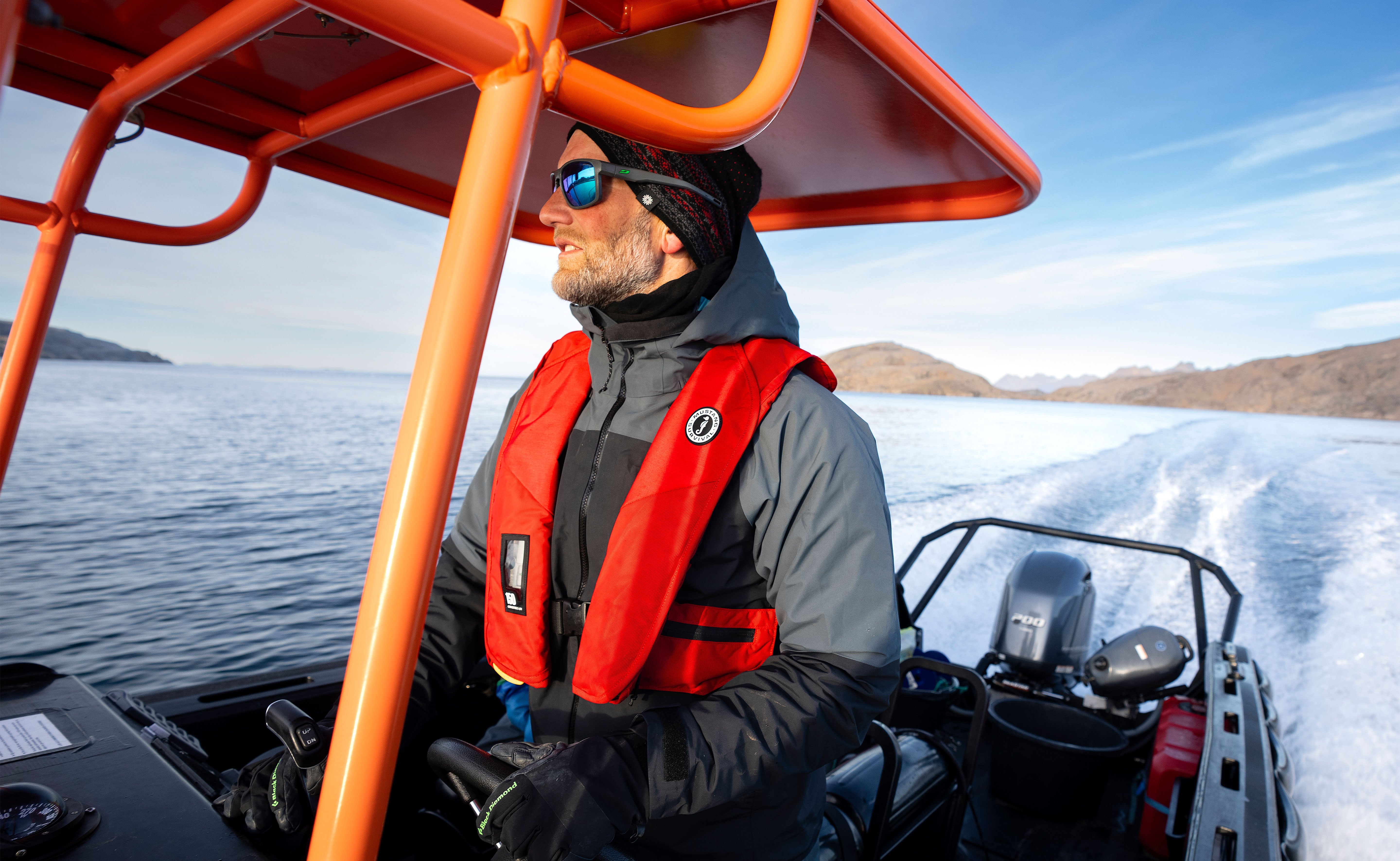 Side profile of a man steering an orange-framed boat while wearing a knit beanie, sunglasses, a grey technical jacket, and a red Mustang inflatable life jacket.