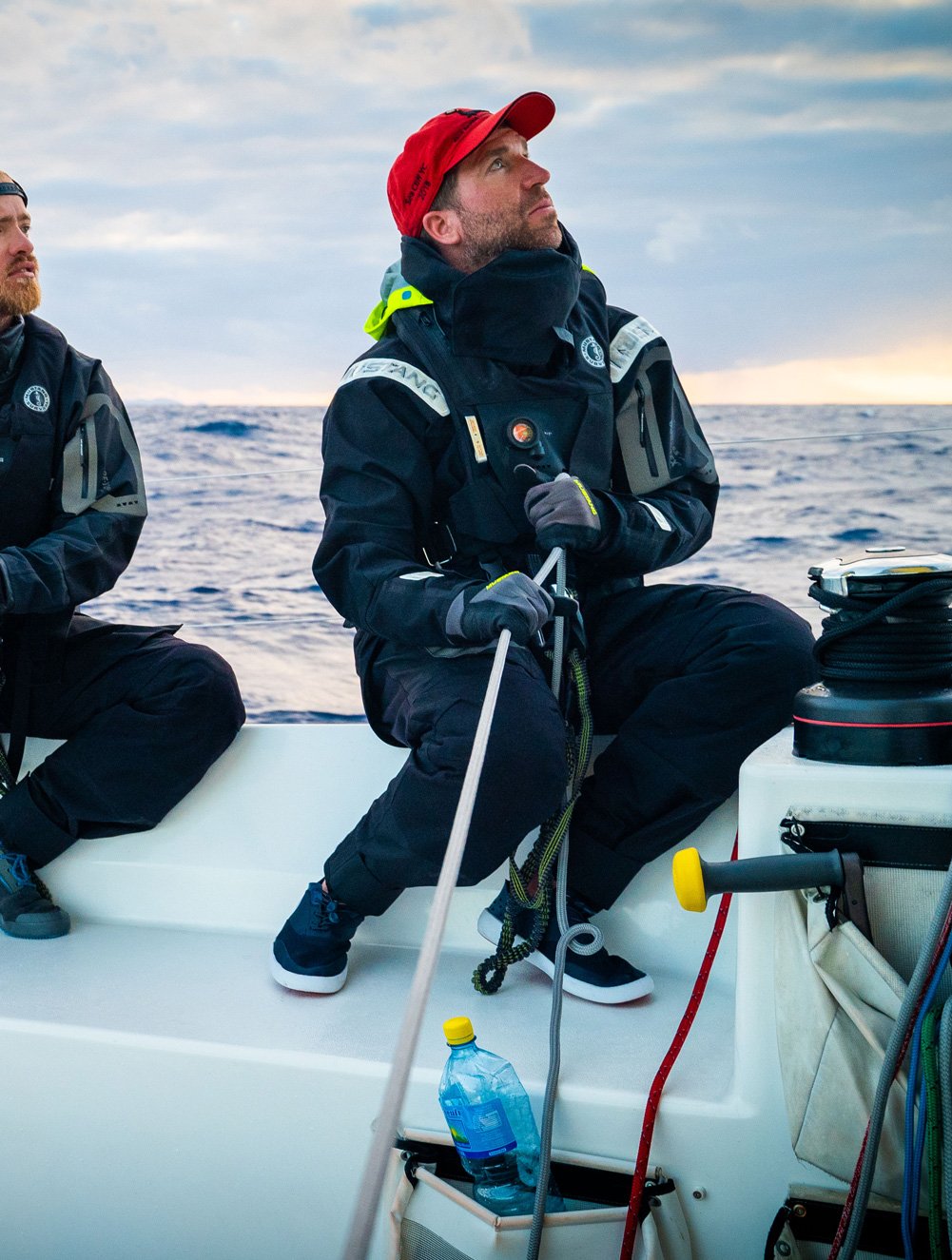 A sailor in a red cap operating lines on a racing yacht at sea, wearing a heavy black Mustang Survival offshore jacket with high-visibility shoulder panels and a raised protective neck collar.