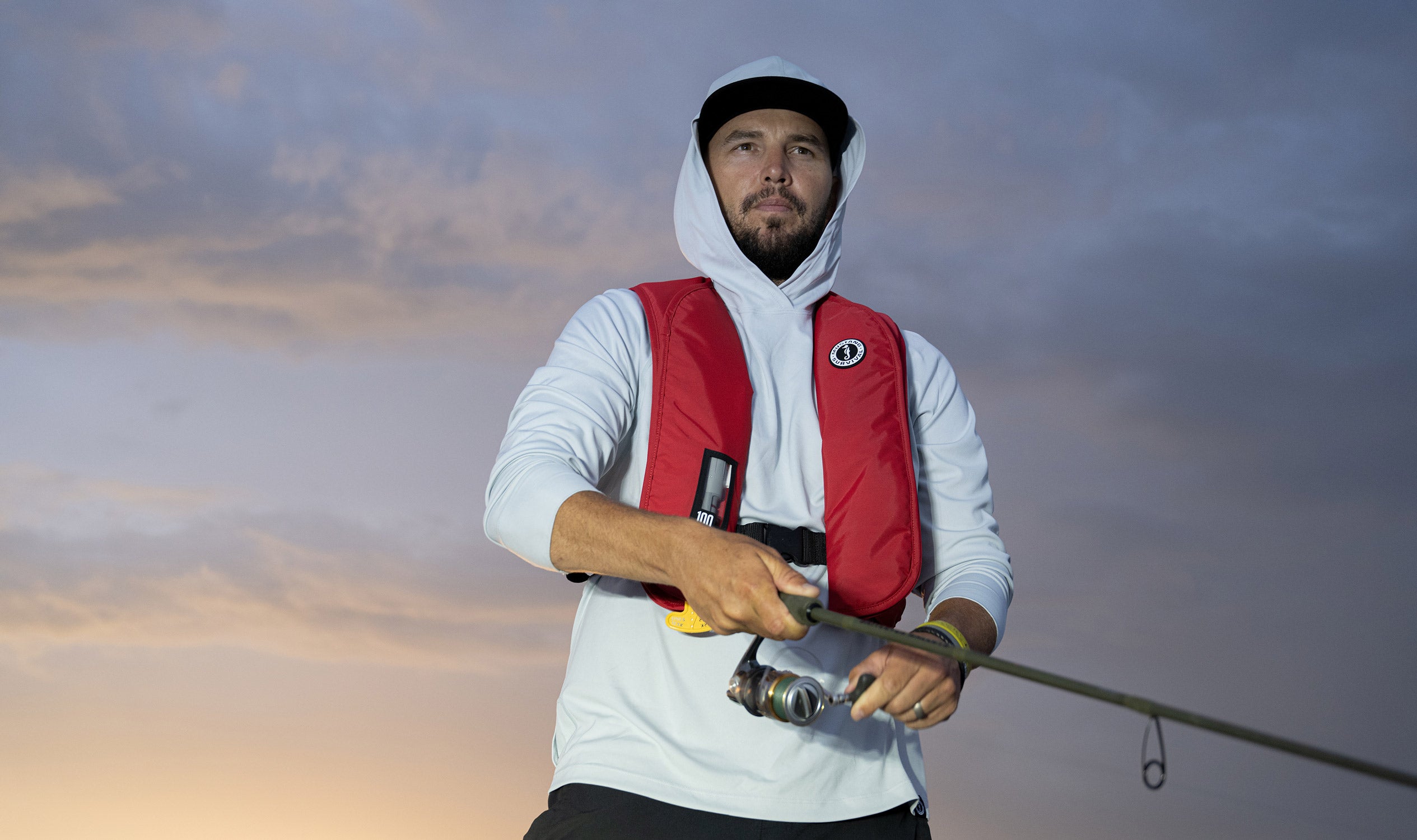 A man fishing from a boat at dusk, wearing a light grey hooded sun shirt and a red Mustang Survival MIT 100 inflatable life jacket.