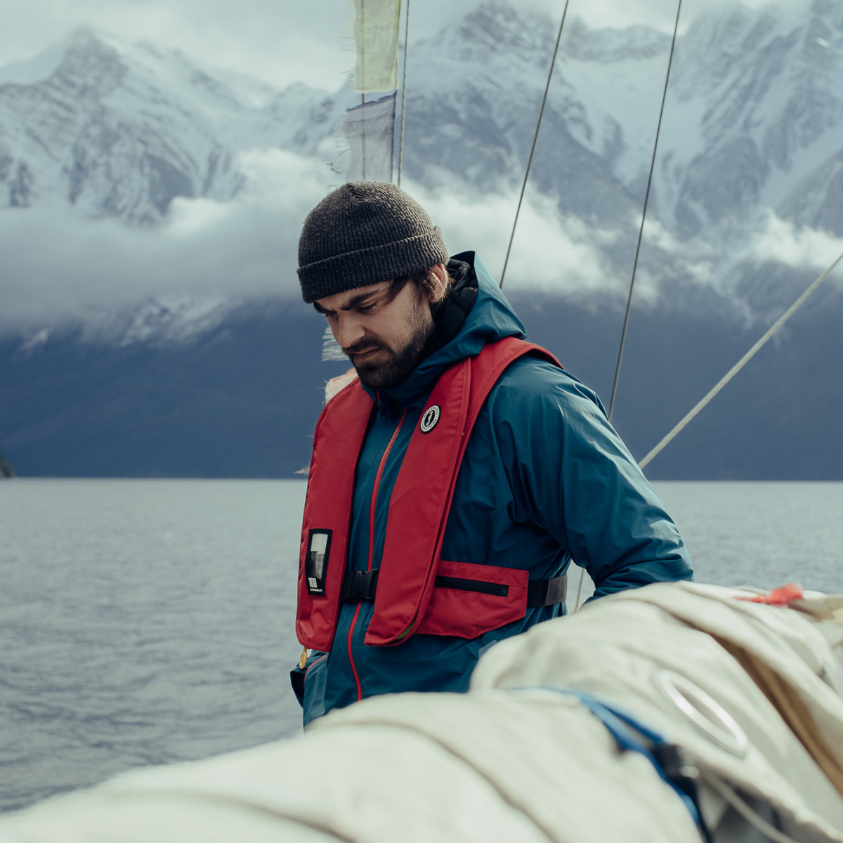 A bearded man on a boat wearing a knit beanie, a blue Mustang Survival Callan waterproof jacket, and a red inflatable life jacket against a background of snow-capped mountains.