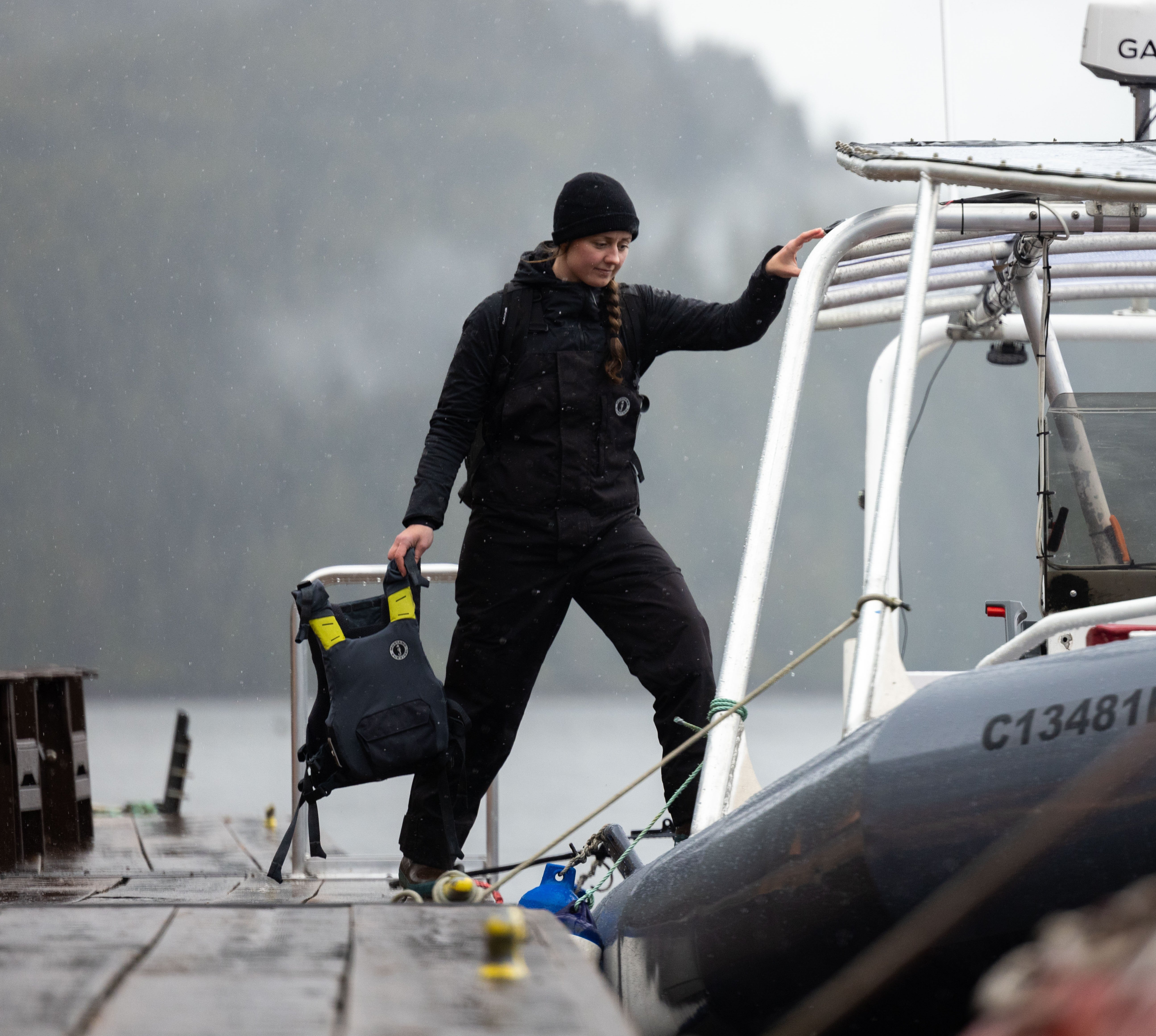 A woman wearing a black beanie and black Mustang Survival flotation bib pants steps onto a boat from a wooden dock while holding a grey and yellow Khymera dual flotation PFD.