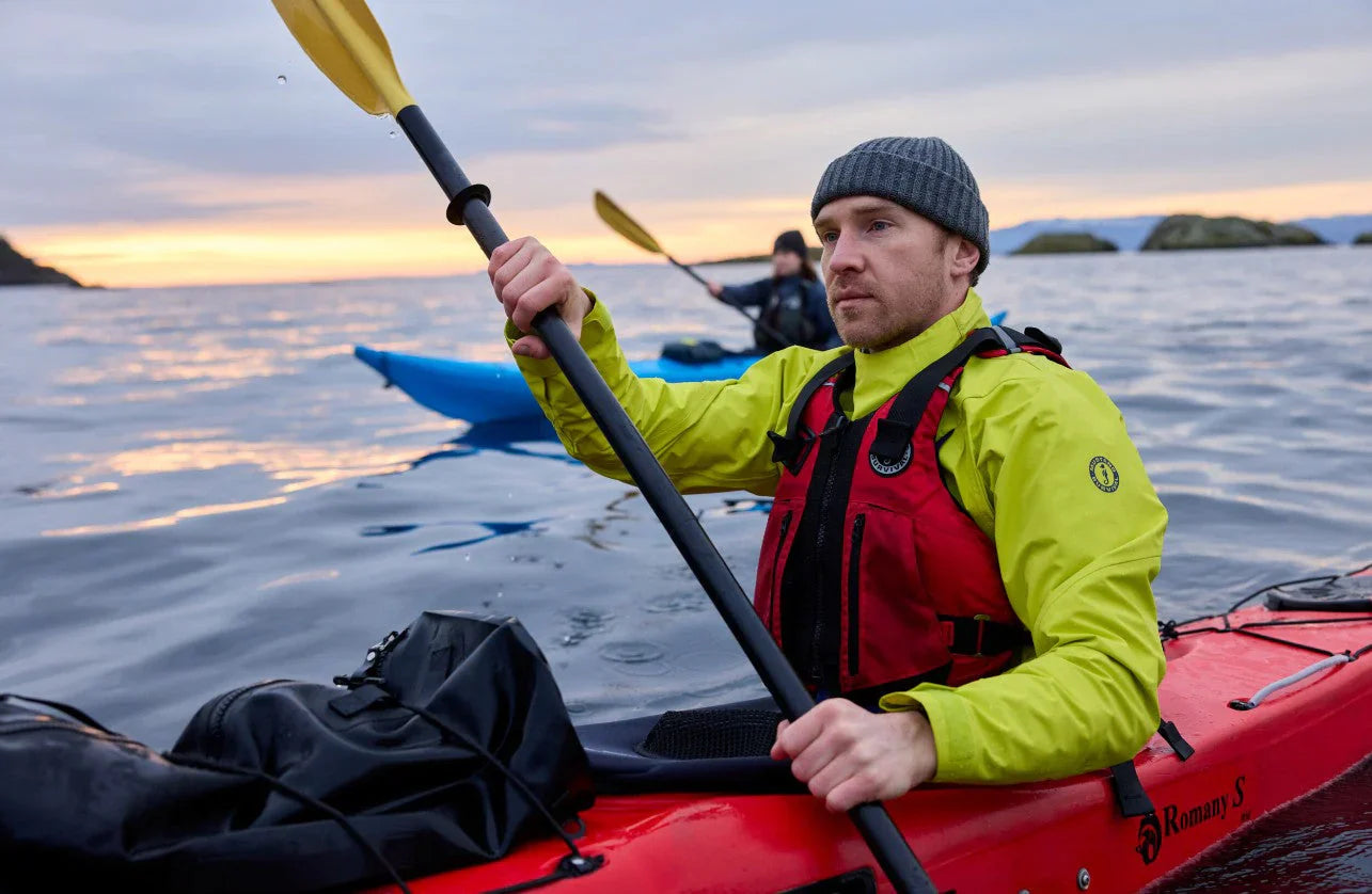 A kayaker wearing a gray Mustang Survival toque and a Neptune and Mahi Yellow Taku Dry Top rows his kayak on cold ocean water at dawn.