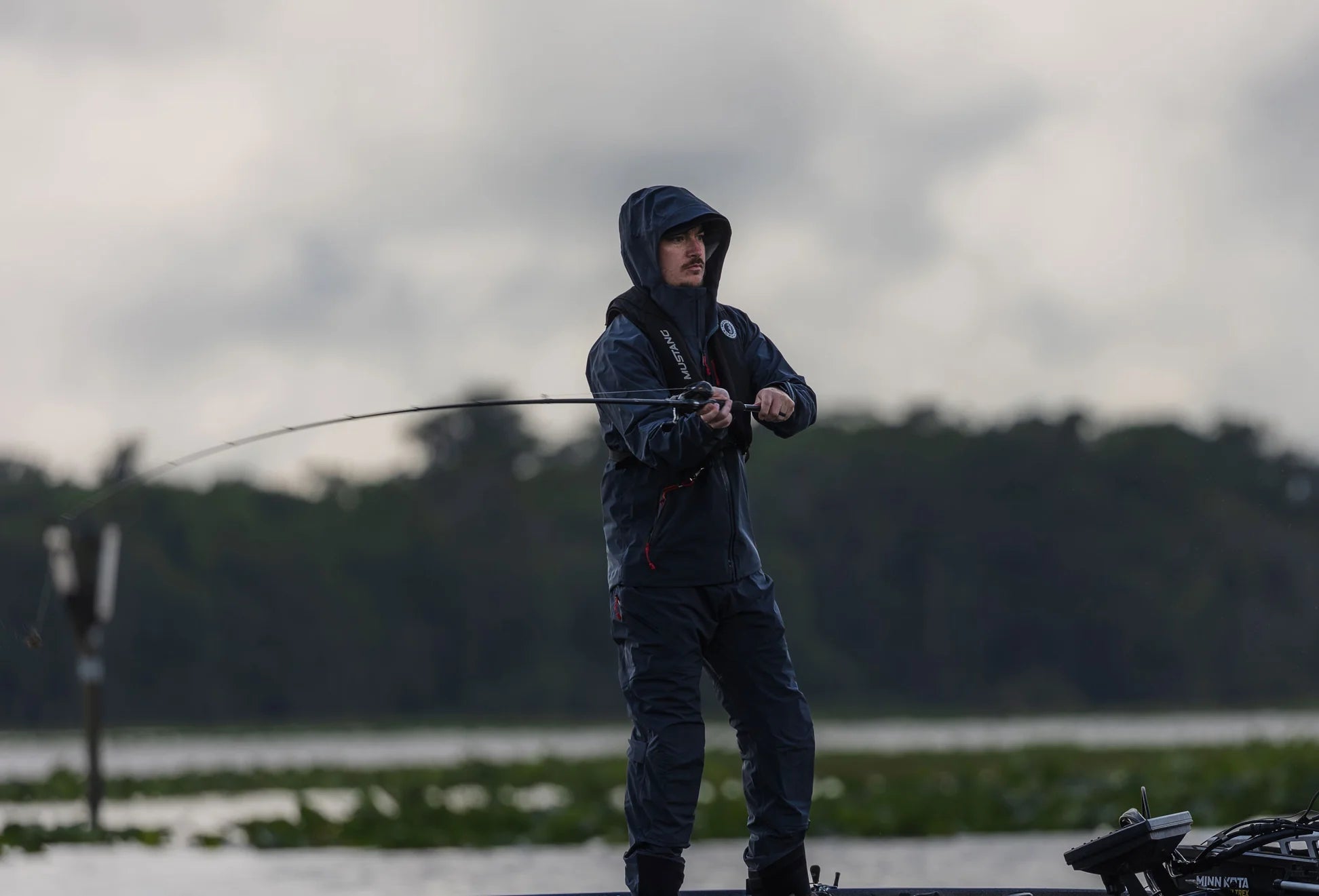 Person wearing a Mustang Survival jacket and life jacket with hood, fishing on a lake from a boat in overcast weather, with forested shoreline in the background.