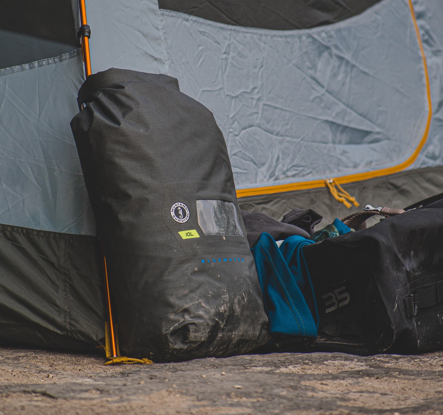 A black Mustang Survival roll-top dry bag leans against a camping tent next to other gear on the ground.
