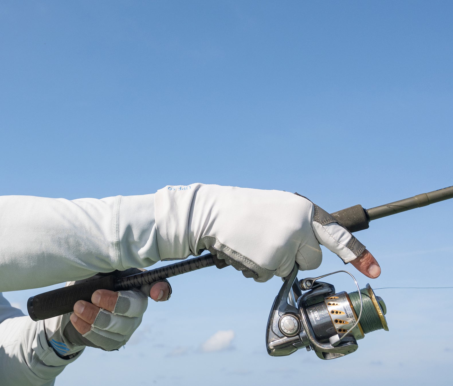 A close-up of a person wearing light grey Mustang Survival UV traction gloves while holding a fishing rod and reel.