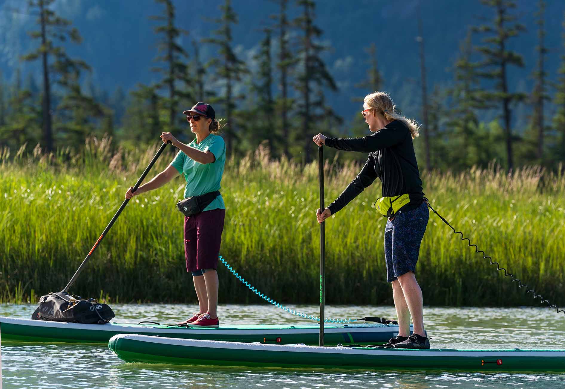 Two women stand-up paddleboarding on a calm river near grassy banks while wearing Mustang Survival inflatable belt pack PFDs.