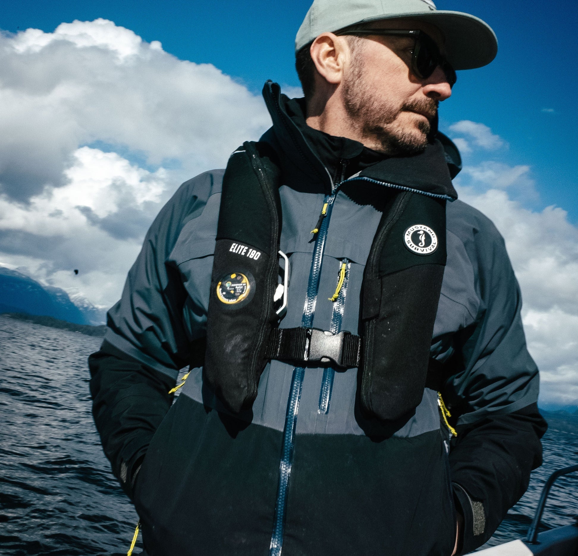 A profile shot of a man on a boat wearing sunglasses, a baseball cap, a technical waterproof jacket, and a black Mustang Survival Elite 190 inflatable PFD.