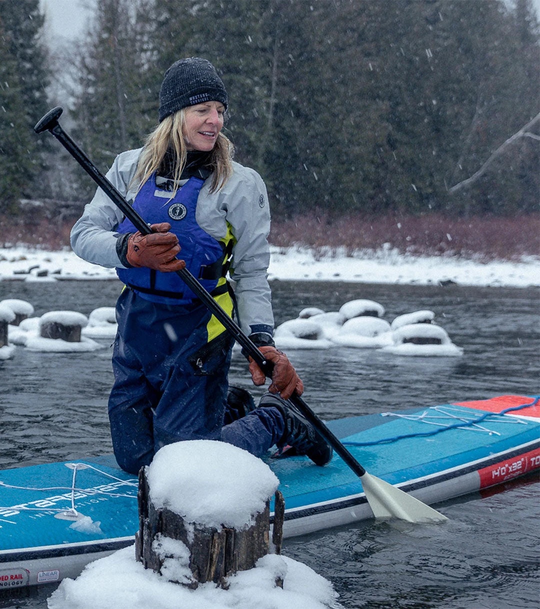 A female paddler wearing a black toque, the grey and blue Women's Helix CCS Dry Suit, and a blue Cascade Foam Vest kneels on her standup paddleboard in a snowy river.