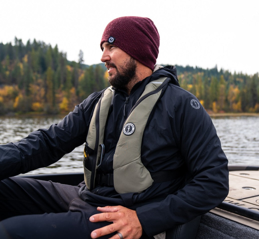 A pro paddler wearing a maroon beanie, black jacket, and an olive green Mustang Survival MIT 100 inflatable PFD smiles while sitting on a watercraft.