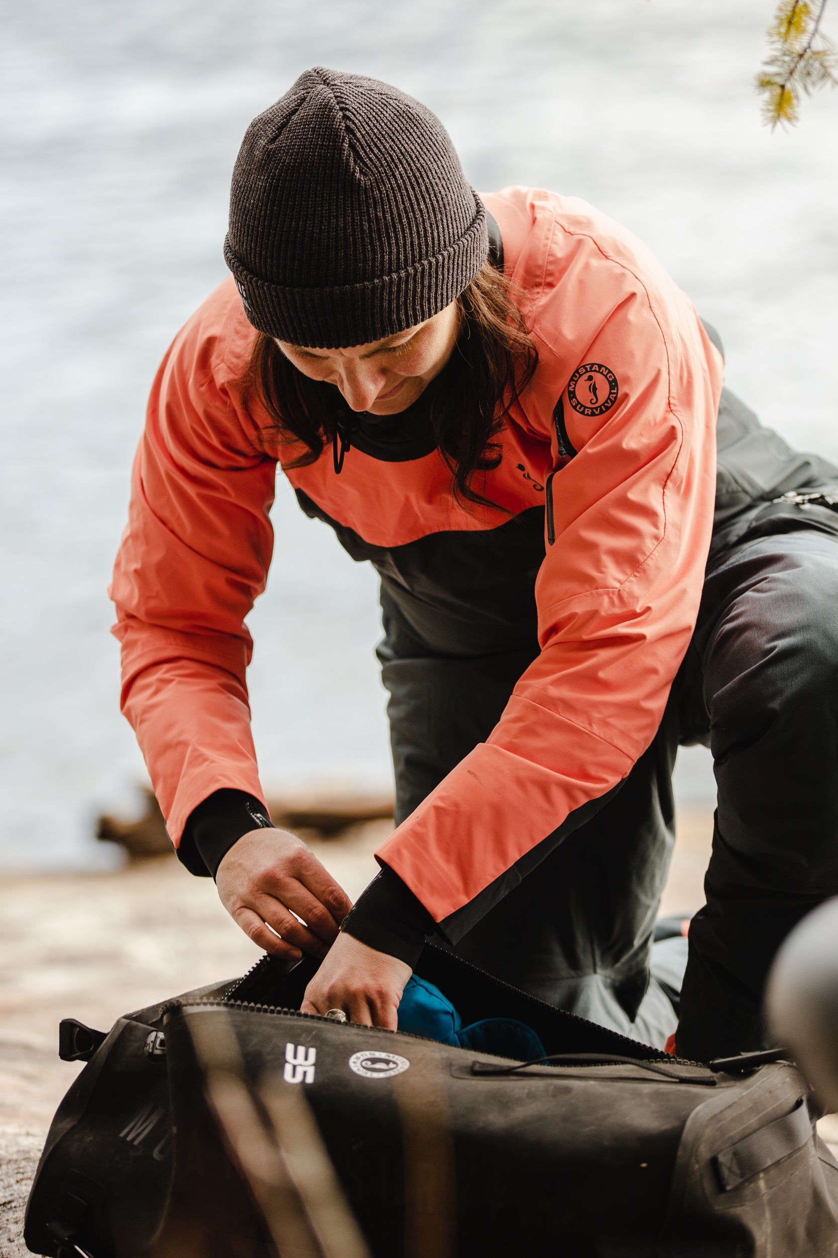 A person in a coral and grey Mustang Survival Helix drysuit kneels to pack a waterproof duffel bag by the water.