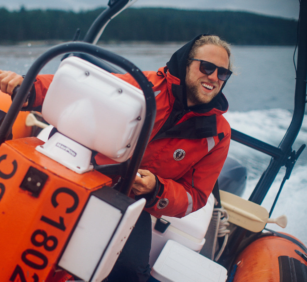 A smiling man steering an inflatable boat through open water while wearing sunglasses and a red Mustang Survival flotation coat with an integrated black hood and collar.