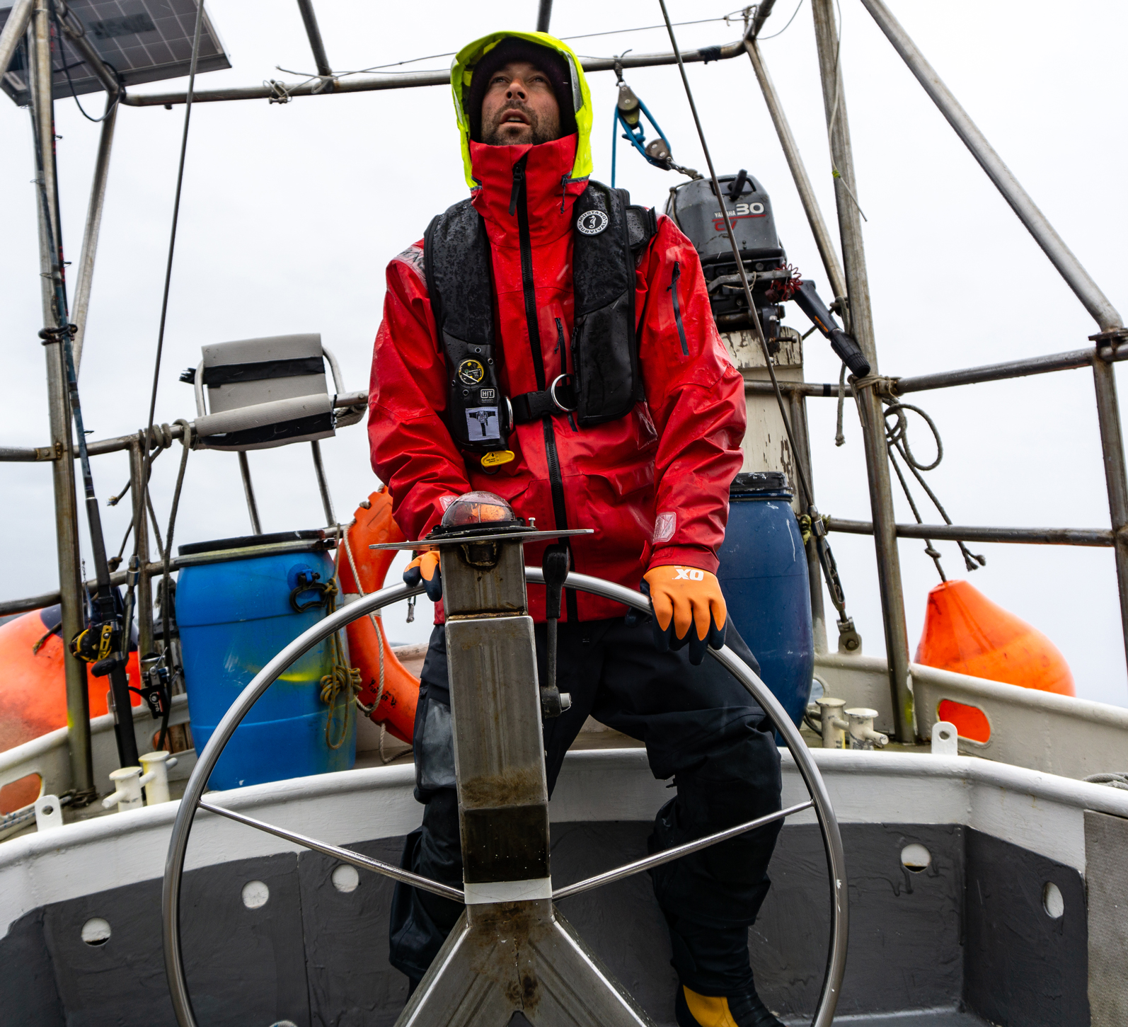 A sailor at a boat's steering wheel wearing a red foul weather jacket with a high-vis hood and a black inflatable life jacket.