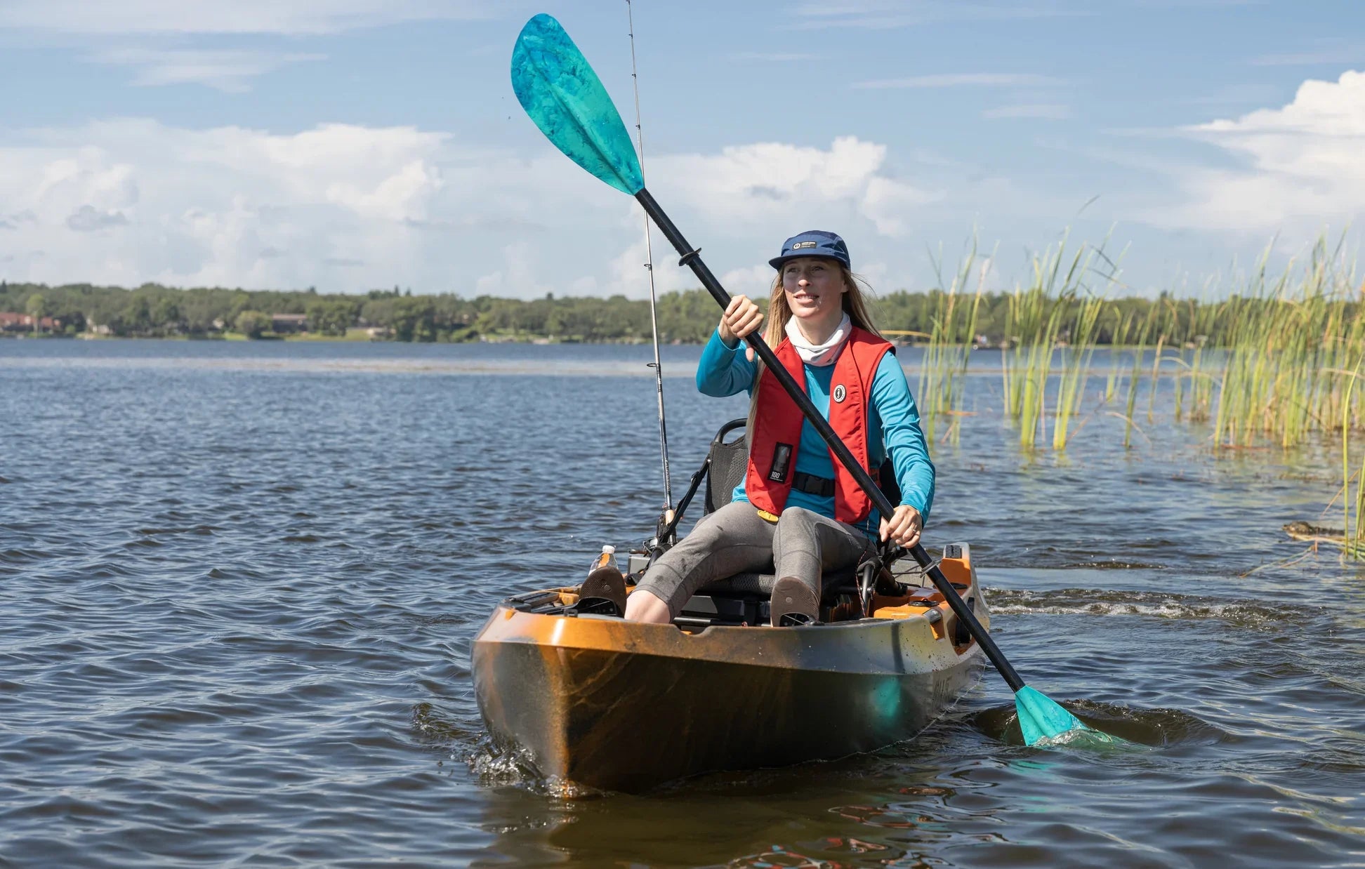 A female angler wearing the bluefin Women's Adelphi UV Long Sleeve shirt and a red MIT 100 Convertible A/M Inflatable PFD paddles her angling kayak.