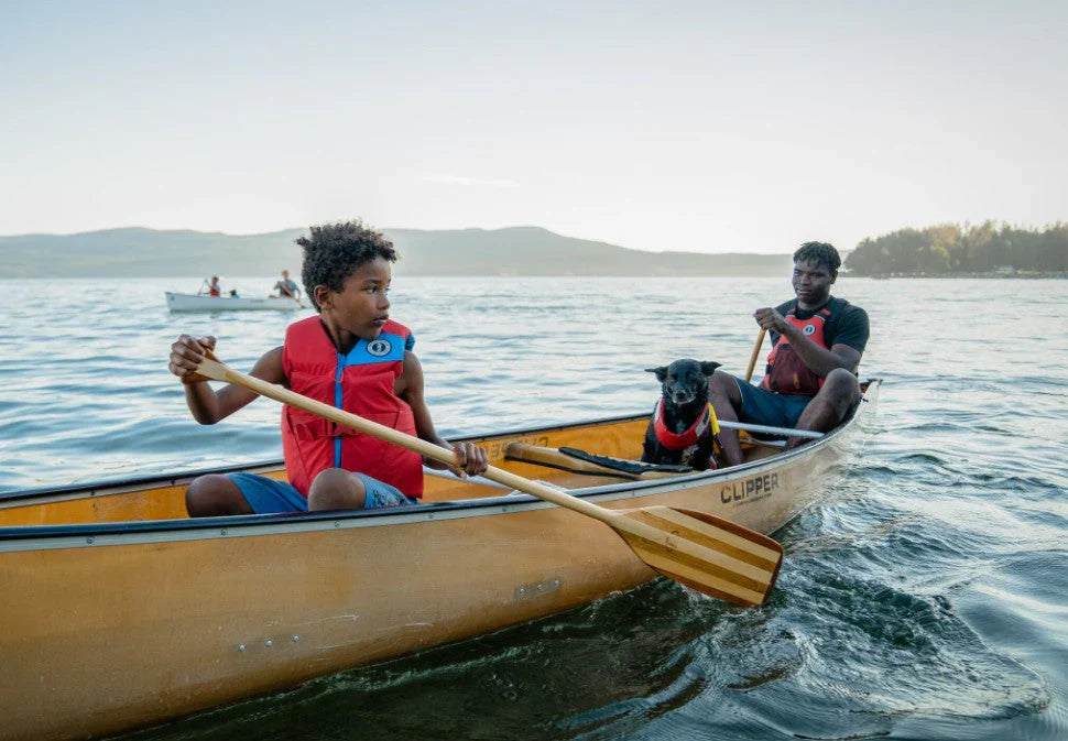 A child in a red Youth Rev Foam Vest and a man in a red Vibe Foam Vest paddle a canoe while the family dog wears the UnderDog Foam Flotation.