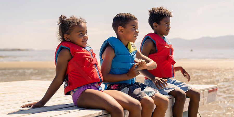 Three children sitting in a row on a wooden dock by the ocean, smiling and wearing blue and red Mustang Survival youth foam life vests.