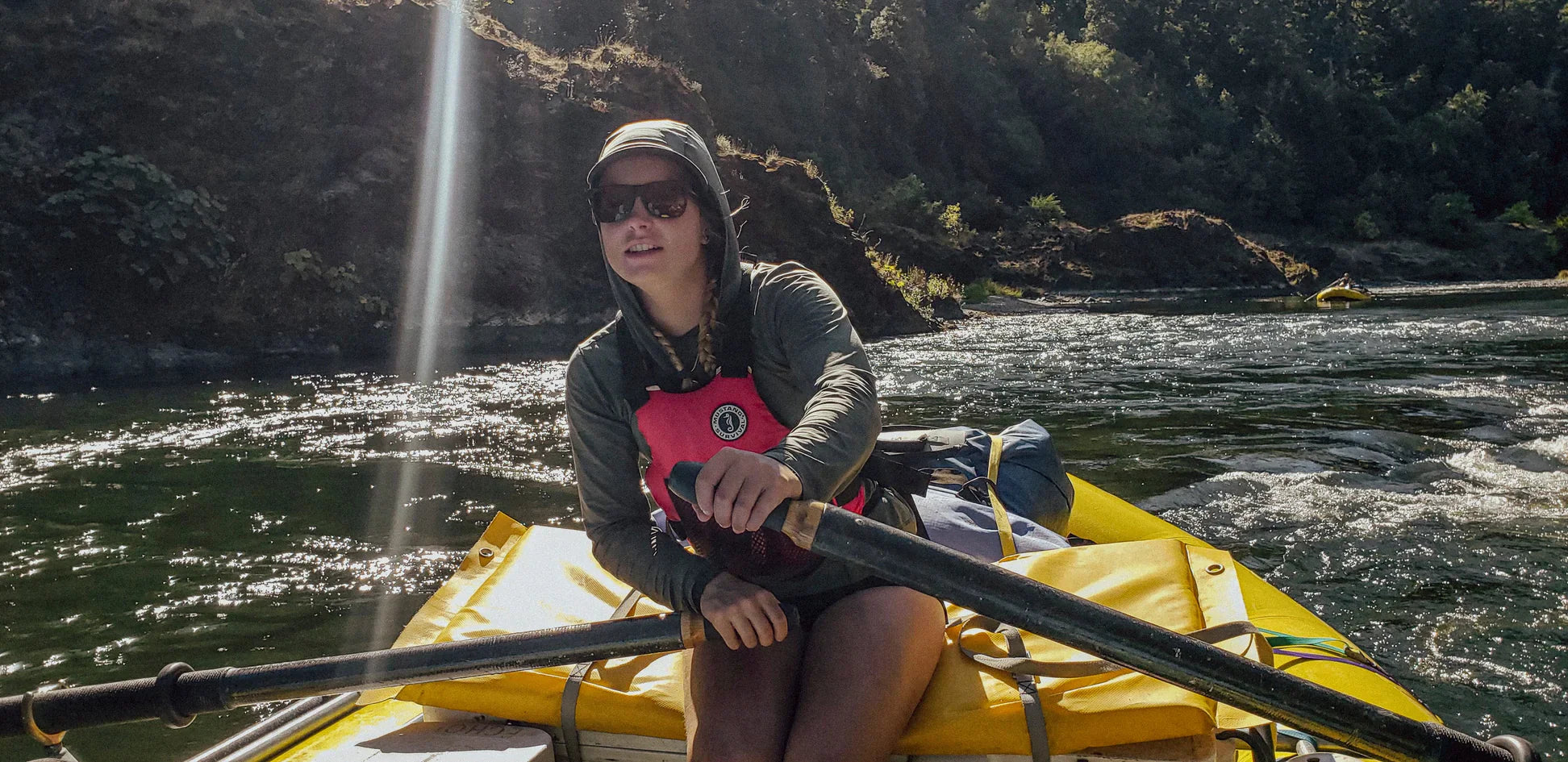 A person wearing a red foam life vest, sunglasses, and a hooded long-sleeve shirt rows a yellow raft down a sunny river bordered by a forested, rocky shoreline.