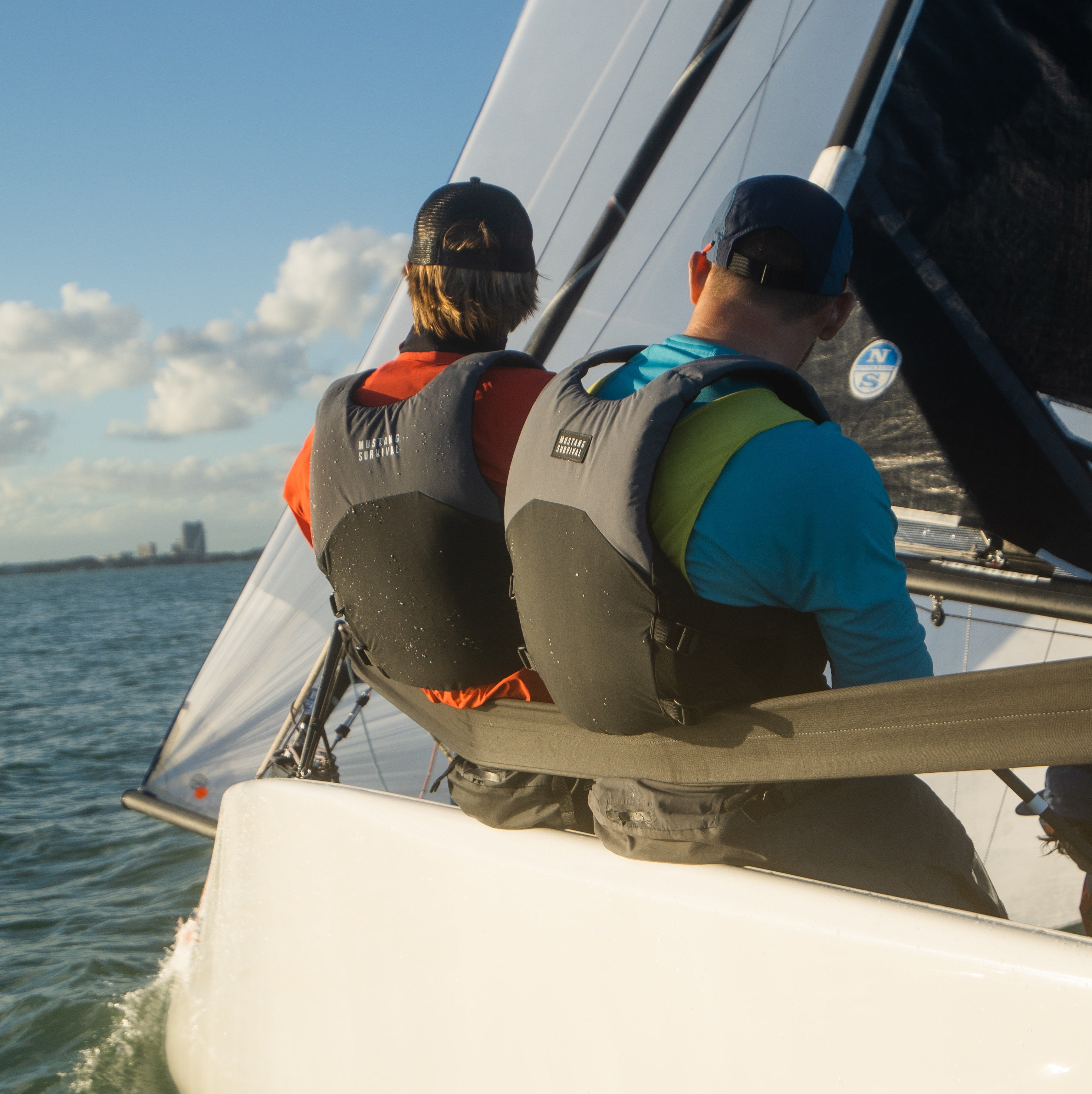 Rear view of two sailors sitting on the edge of a sailboat on sunny water, wearing baseball caps, long sleeve shirts, and grey Mustang PFDs.