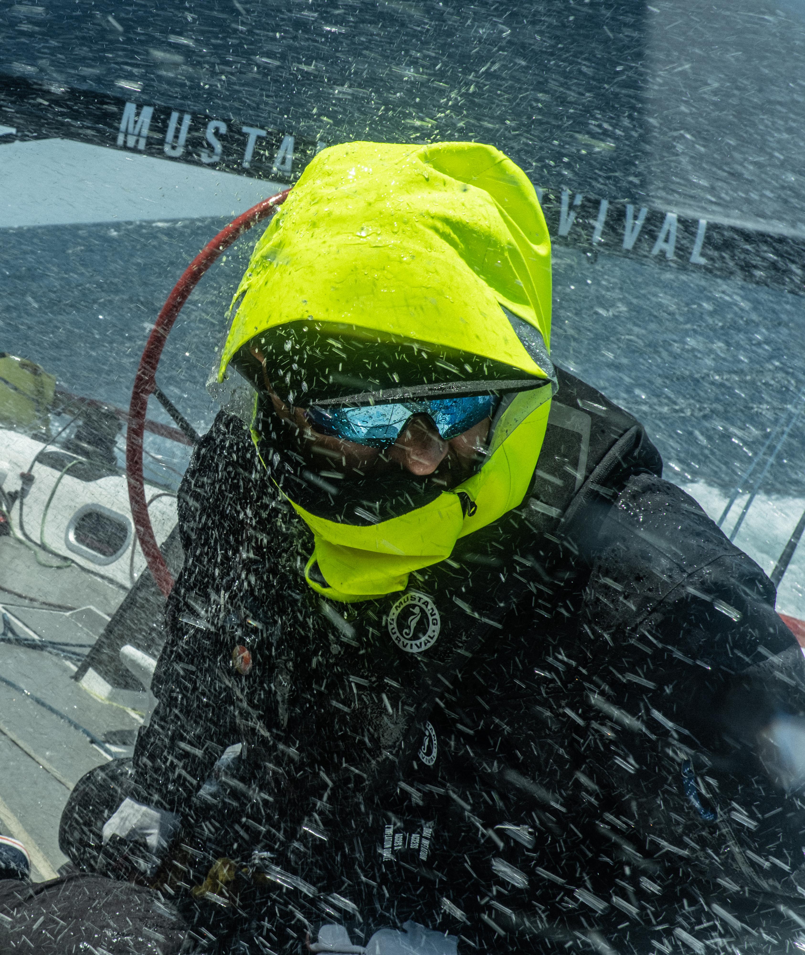 Close-up of a sailor battling heavy ocean spray on a racing boat, wearing a black Mustang Survival EP 6.5 jacket with an integrated high-visibility neon yellow hood and face shield.