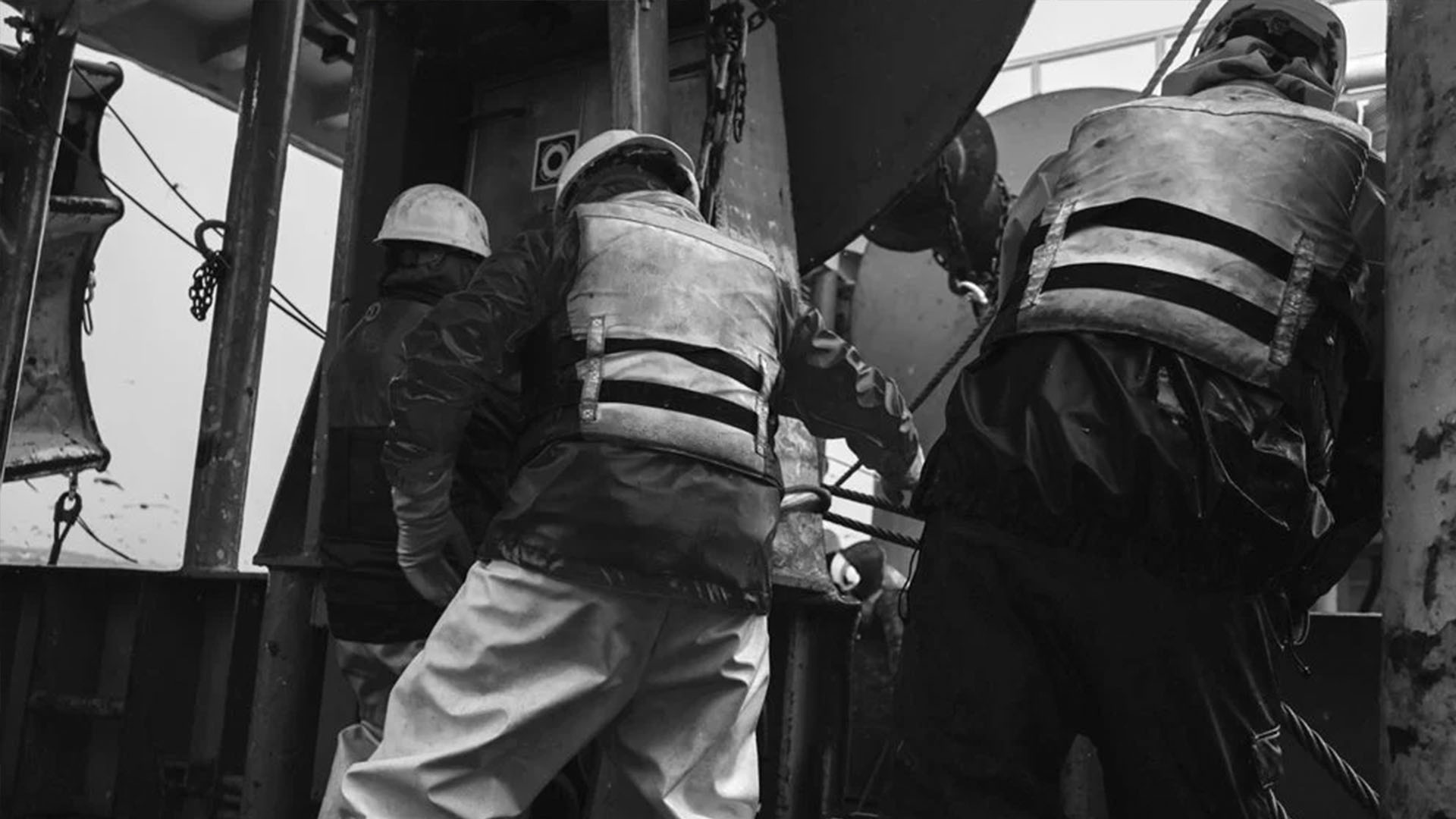 A black and white photo showing three deckhands working on a commercial vessel, wearing hard hats, foul weather gear, and heavy-duty industrial flotation vests with reflective tape.
