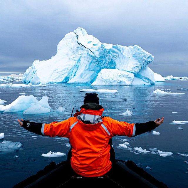 Back view of a person on a boat with arms outstretched toward a large iceberg, wearing an orange Mustang Survival flotation coat.