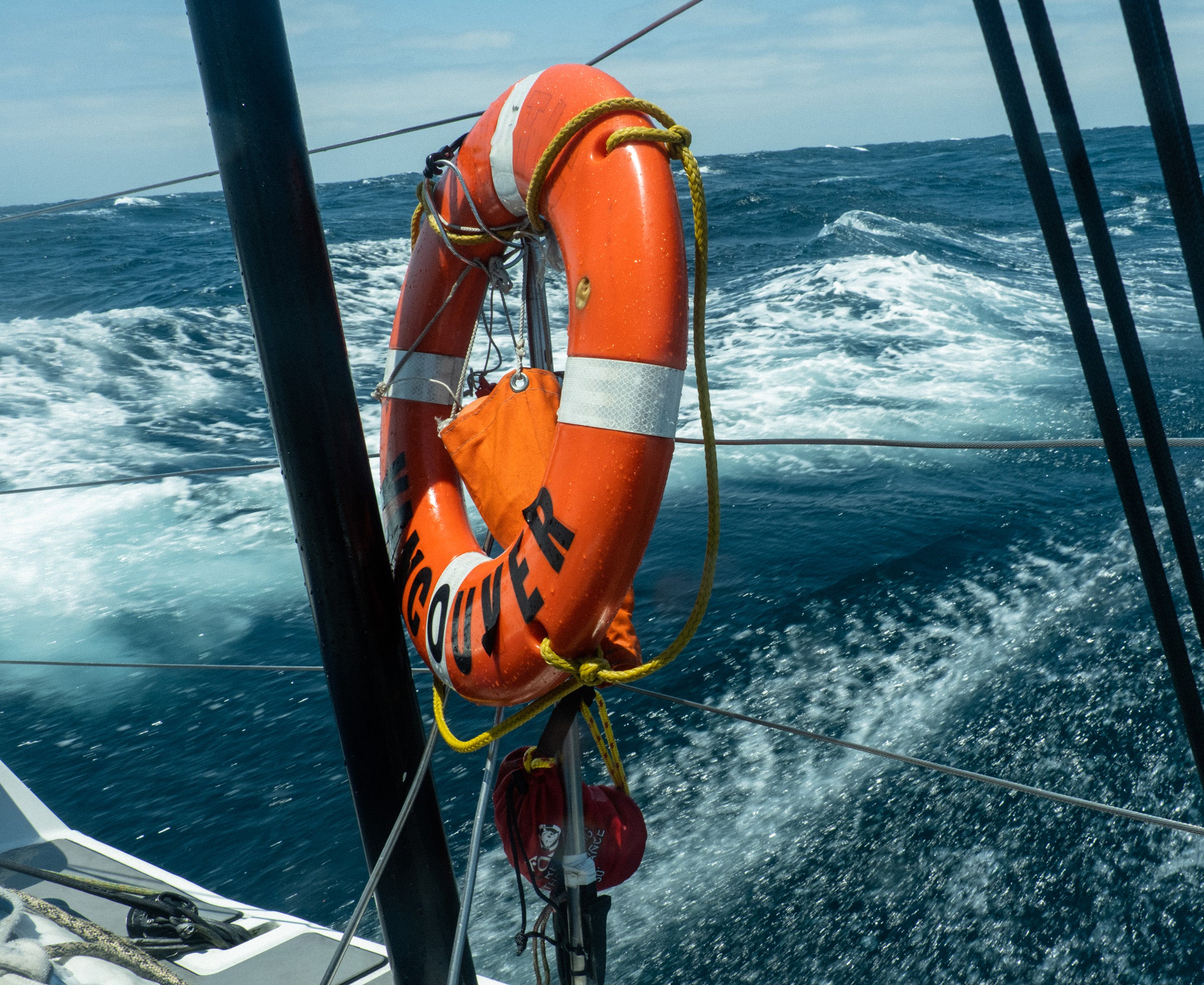 An orange lifebuoy ring with reflective tape mounted on the deck railing of a sailboat moving through rough ocean water.