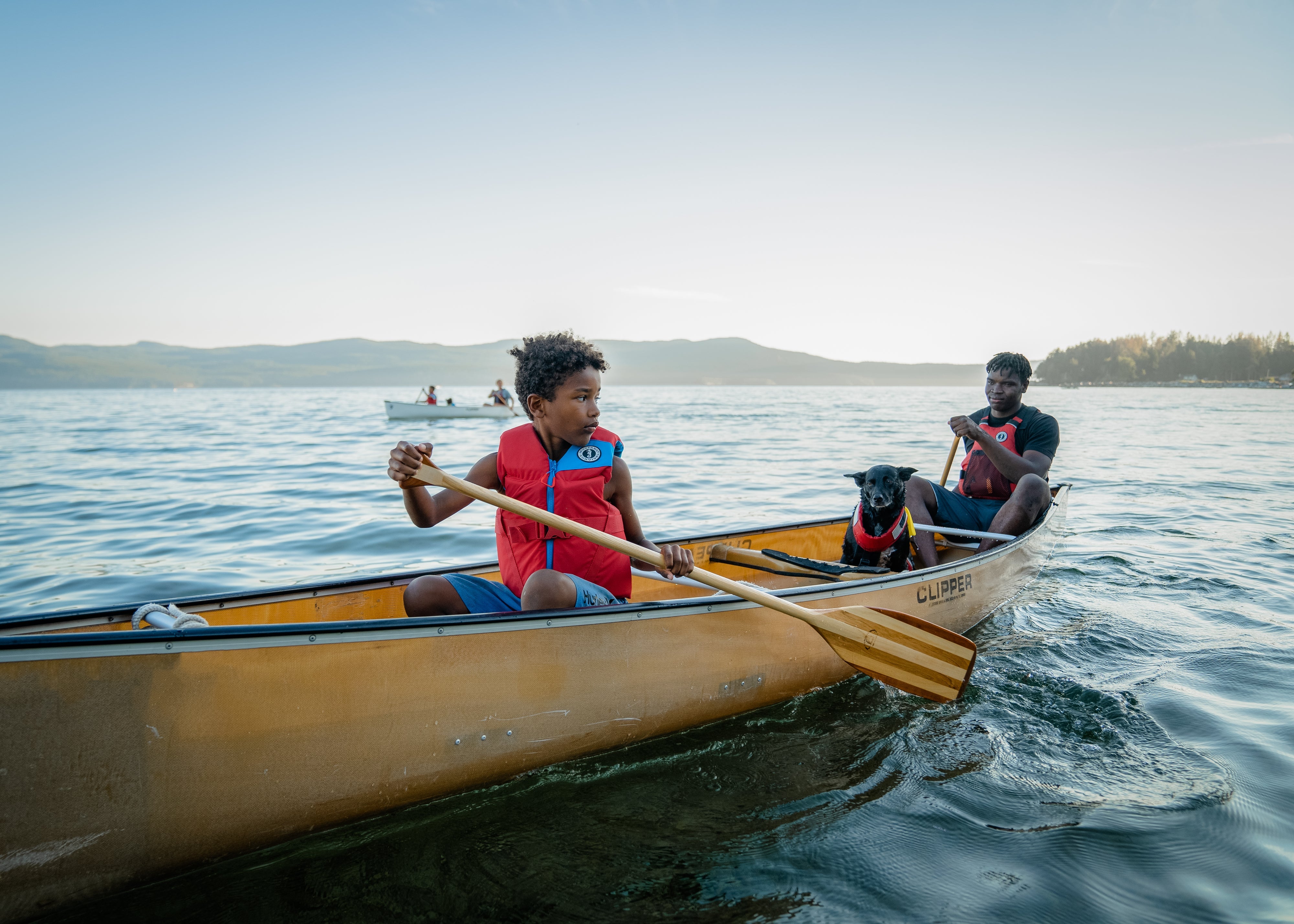 A boy in a red Rev foam life vest paddling a canoe, with an adult and a dog wearing PFDs in the background.