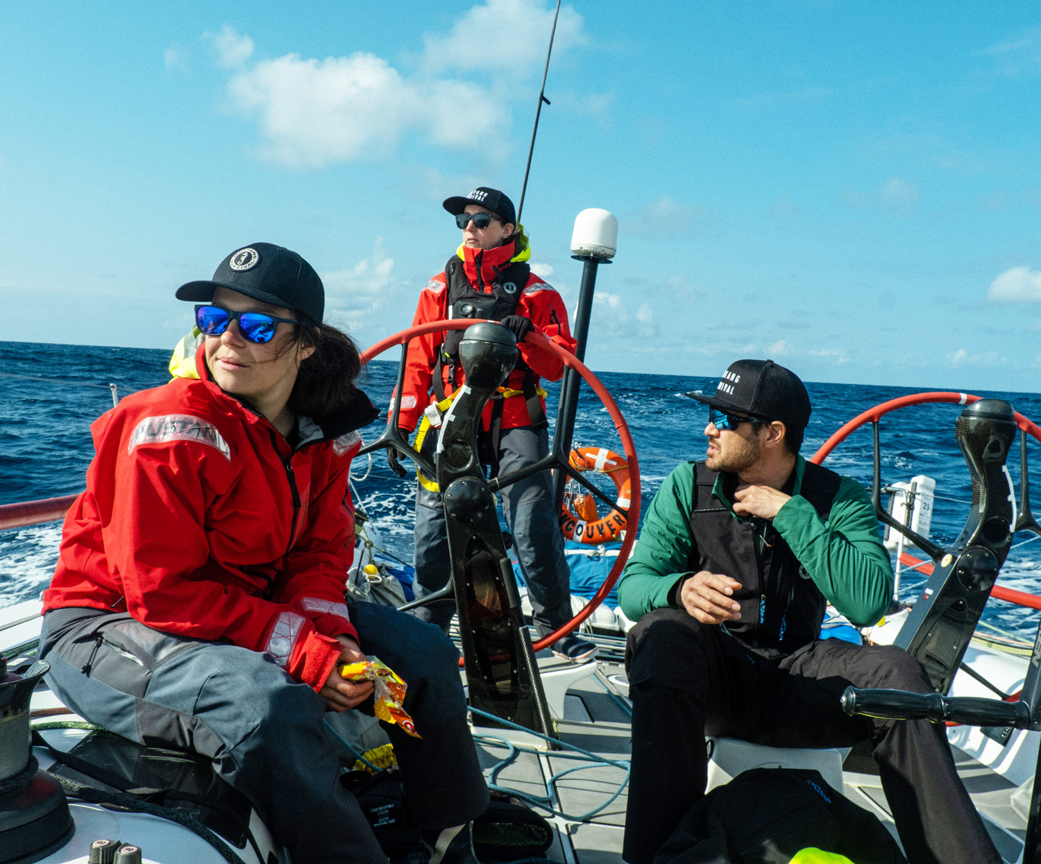 Three pro sailors wearing black hats and offshore gear navigate a sailboat on blue water, with one person at the steering wheel.