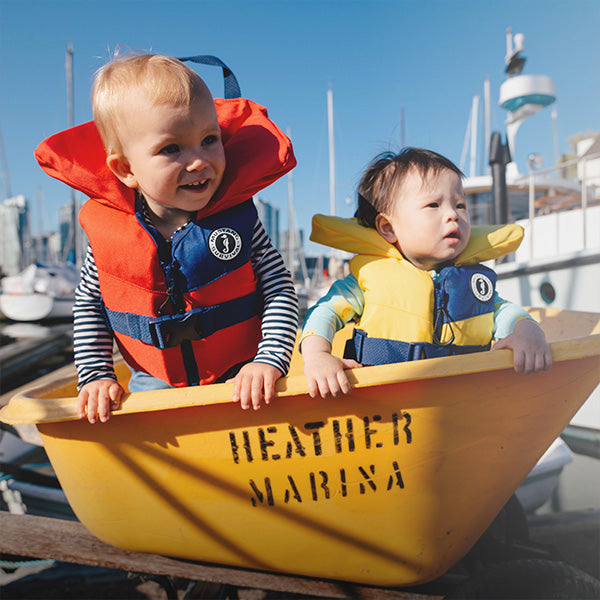 A toddler and a baby sitting together in a yellow tub at a marina, wearing red and yellow Mustang Survival infant Lil Legends foam life vests.