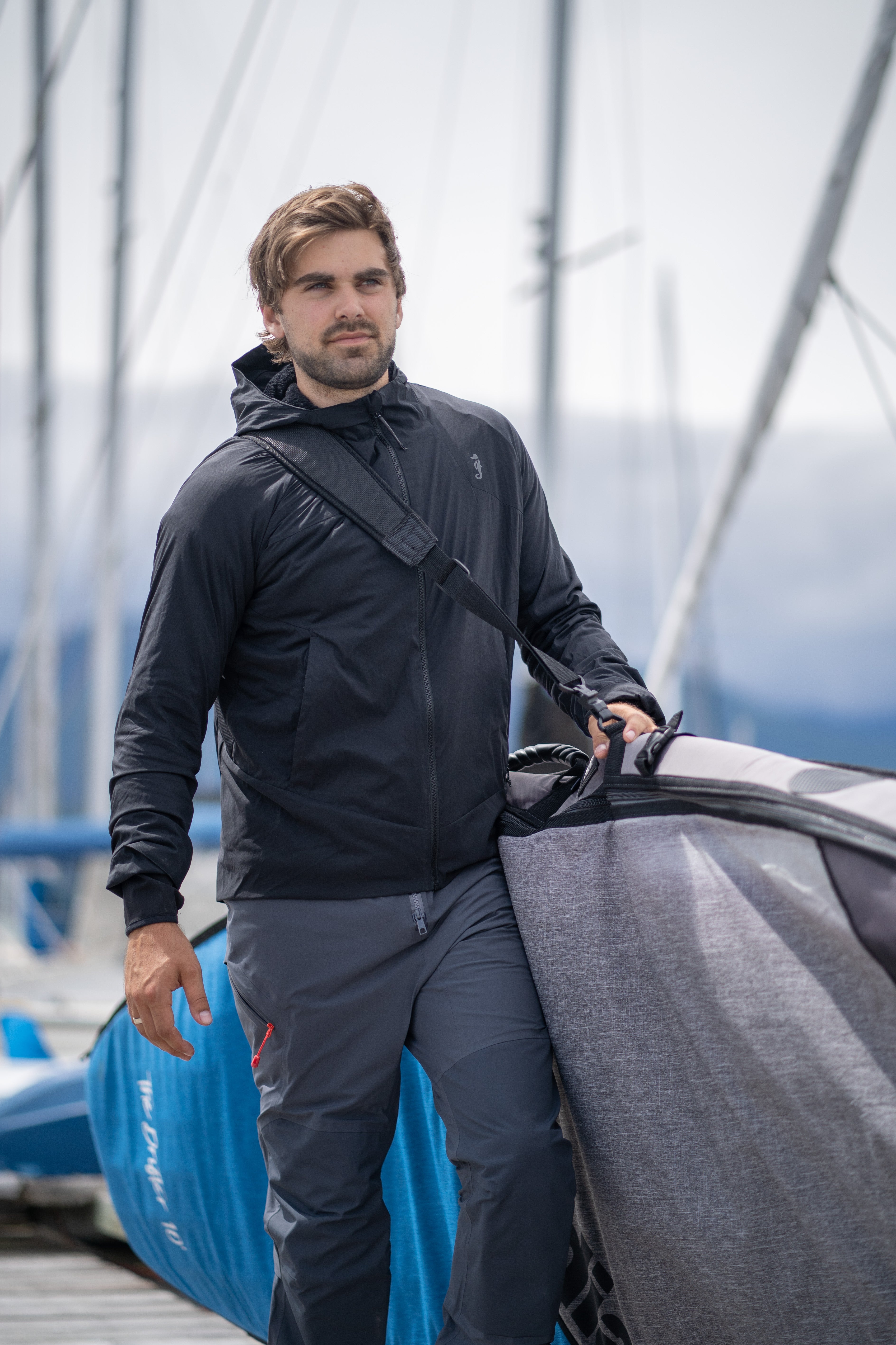 A man carrying a grey foil board bag on a marina dock while wearing a black Mustang Survival hooded jacket and grey technical pants.