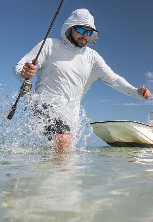 A man wading through clear ocean water with a fishing rod next to a skiff boat, wearing a light grey Mustang Survival UV sun hoodie, sunglasses, and a hat.