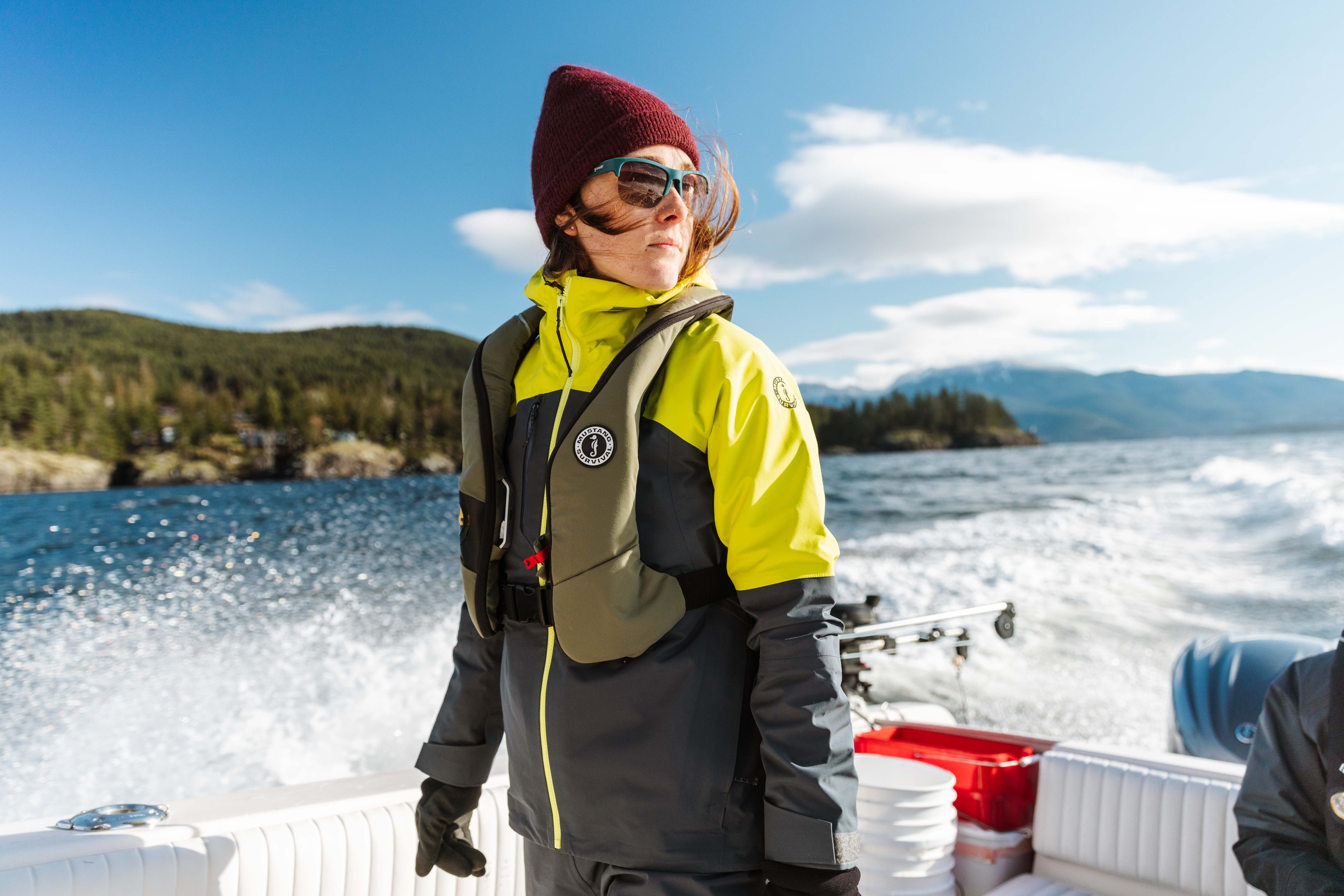 A woman wearing a beanie, sunglasses, and a life vest stands on a moving motorboat with forested mountains in the background.