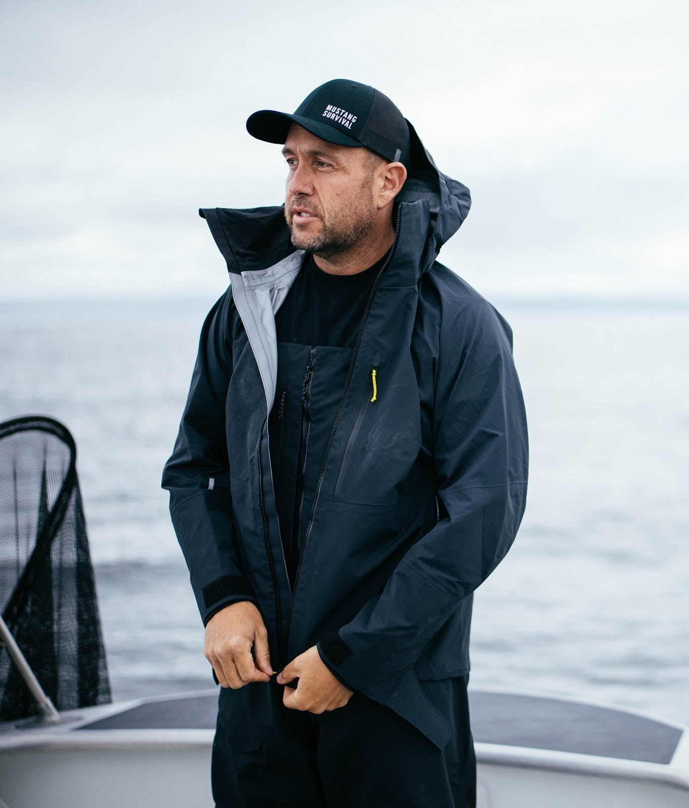 A man on a boat wearing a black Mustang Survival hat, waterproof jacket, and bibs from the men's marine apparel collection.