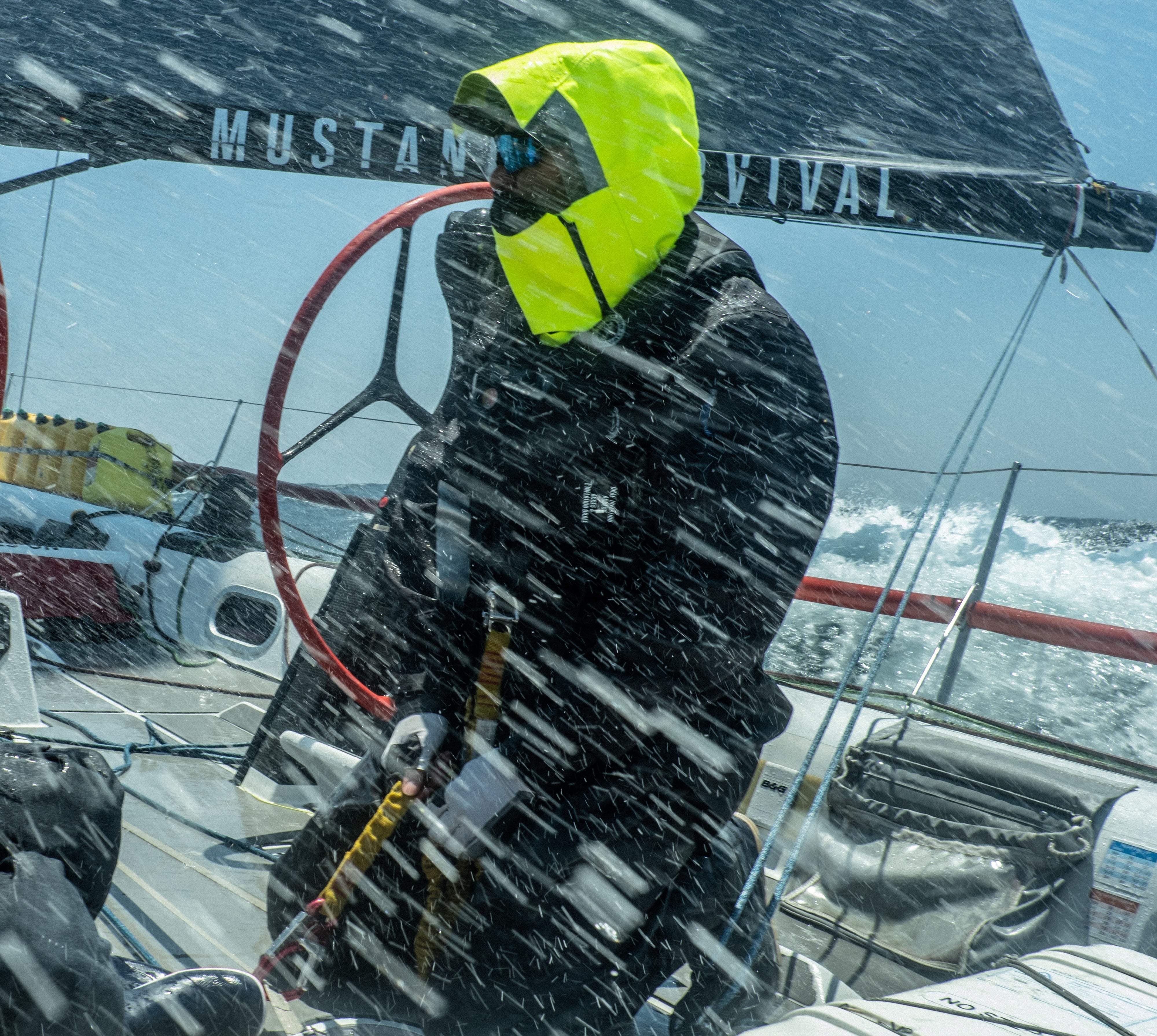 A sailor at the helm of a racing sailboat amidst heavy ocean spray, wearing a black Mustang Survival EP 6.5 Ocean Jacket and Salopette with a high-visibility neon yellow hood and face shield.
