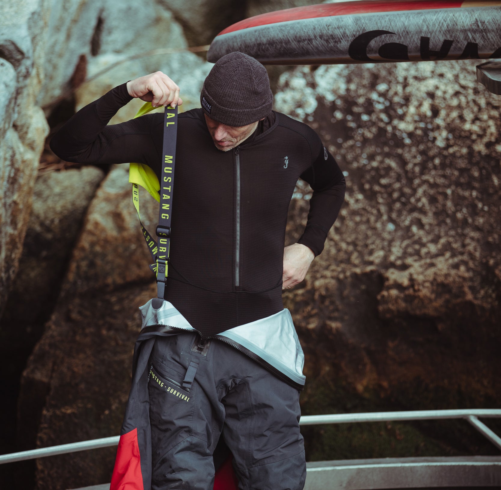 A man adjusting the suspenders of his drysuit while wearing a black Mustang Survival thermal zip-up base layer and a knit beanie hat.