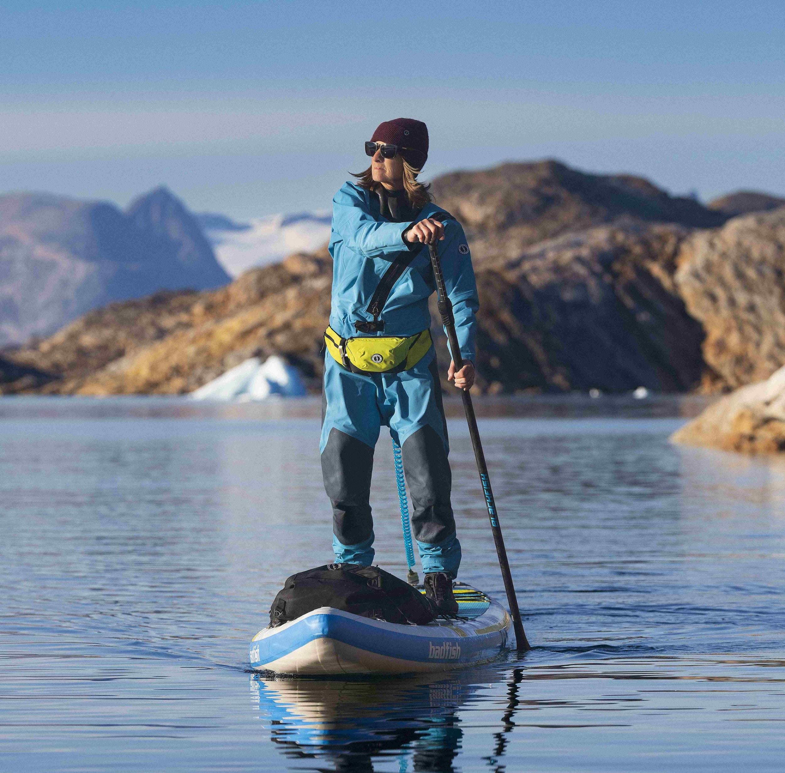 A woman in a blue drysuit and yellow belt pack stands on a paddleboard on calm water with rocky mountains behind.