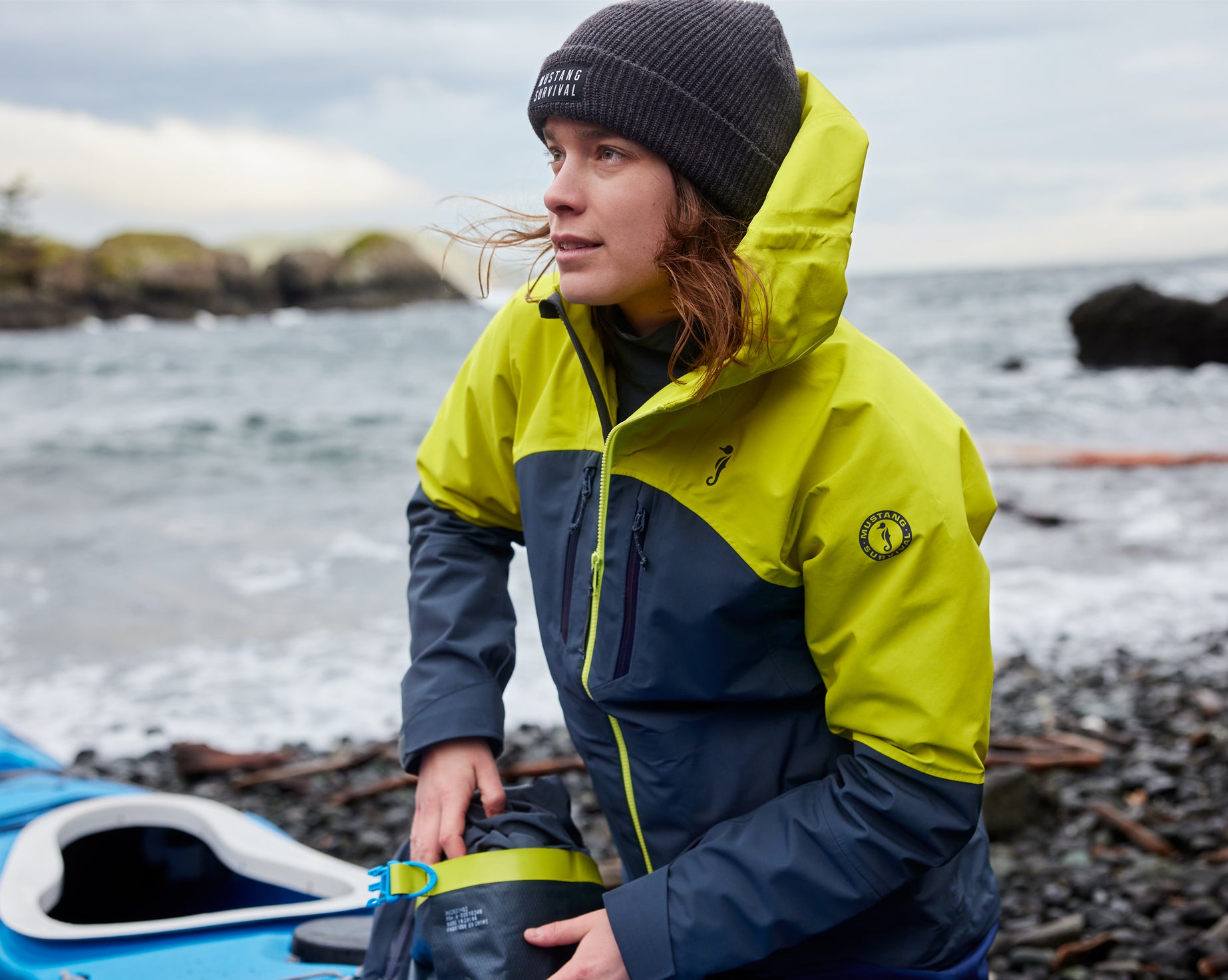A woman wearing a yellow Taku jacket and beanie kneels by a blue kayak on a rocky shore, holding a small dry bag.