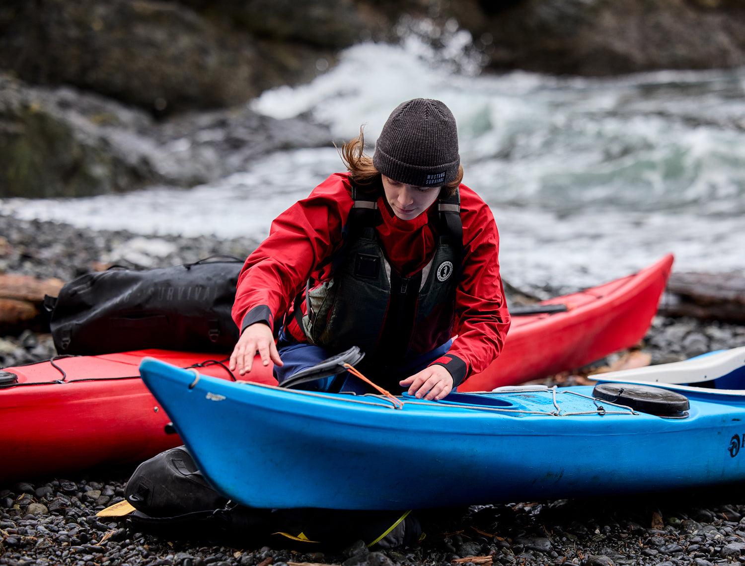 A person wearing a red jacket and beanie prepares a blue kayak on a rocky shore, with a red kayak in the background.