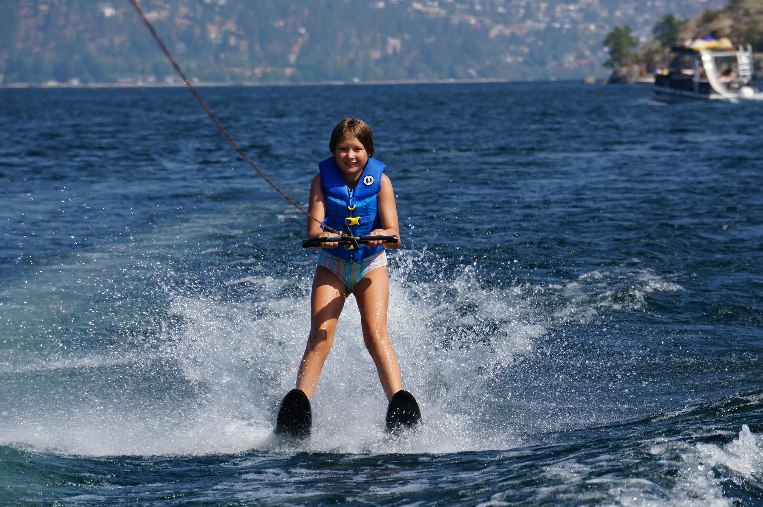 A young girl wearing a blue life jacket smiles while water skiing on a wide, sunny lake, kicking up white spray.