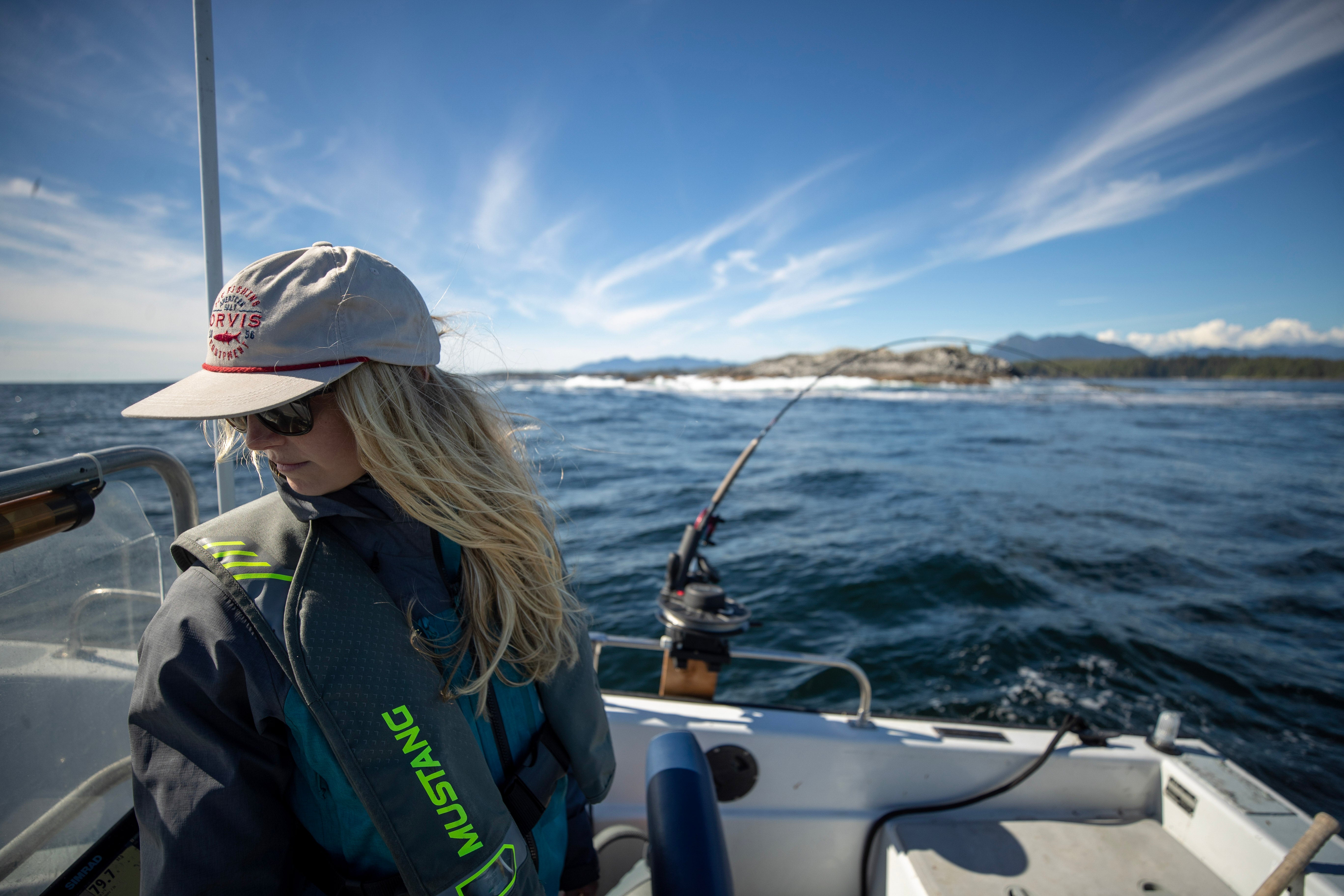 Woman wearing a Mustang Survival life jacket and cap, sitting on a boat during a cold water fishing trip, with a rod set up beside her and ocean, mountains, and blue sky in the background.