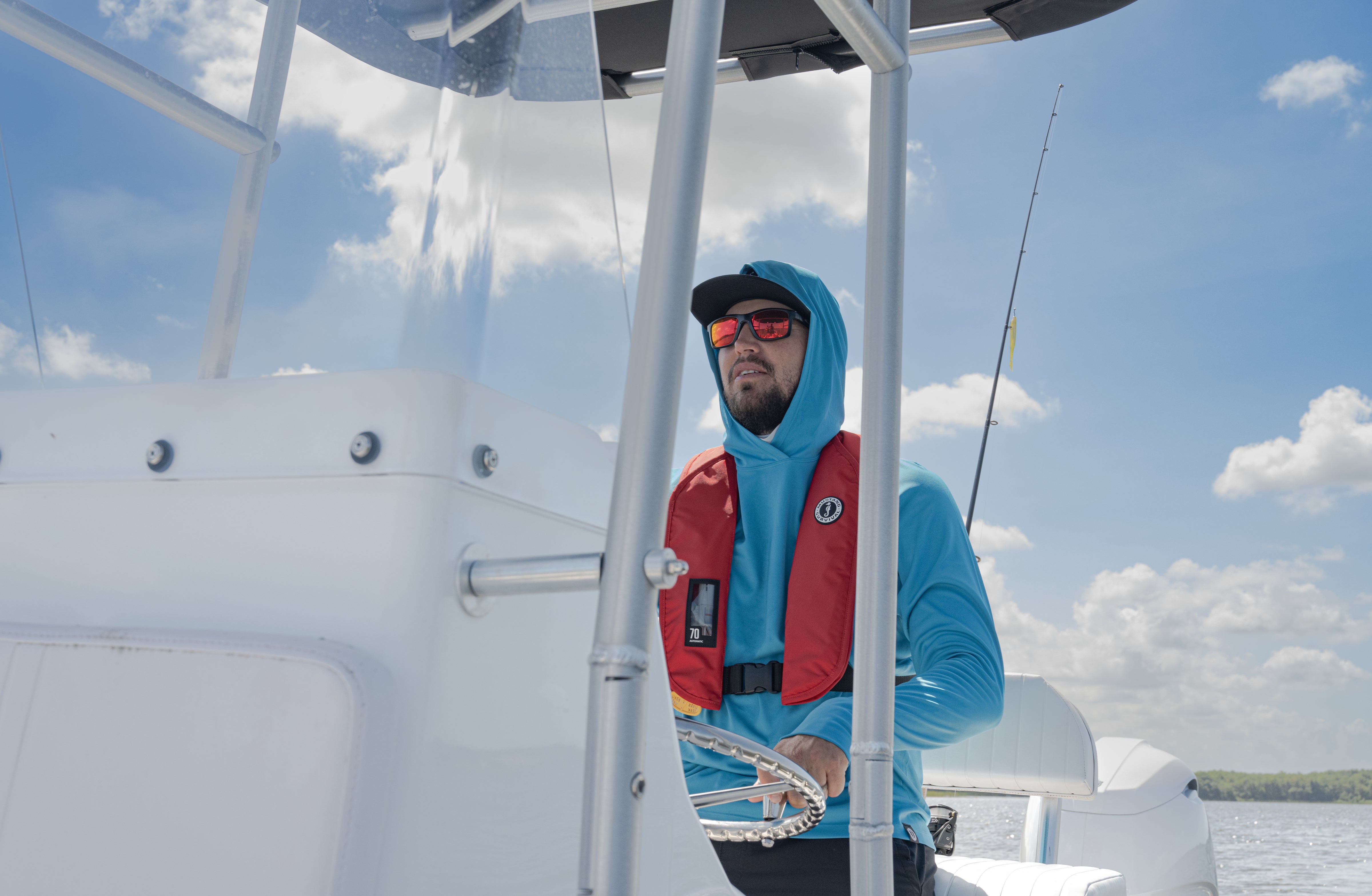 Man wearing a Mustang Survival life jacket and sunglasses, standing on a boat during a warm saltwater offshore fishing trip, with a rod and open ocean in the background under a partly cloudy sky.