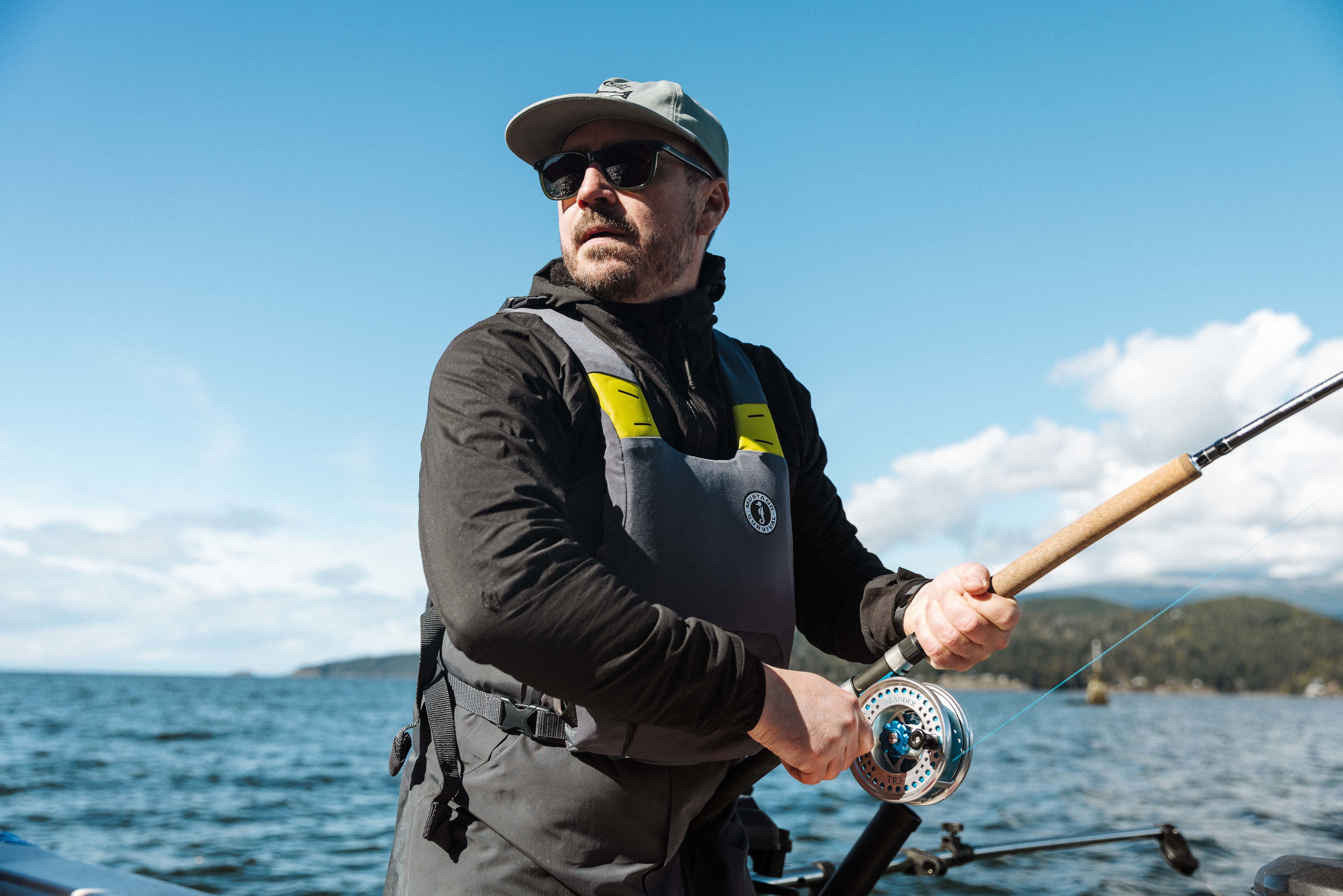 An angler reeling in a fly fishing rod on a boat while wearing sunglasses, a grey hat, and a grey Mustang Survival foam life jacket vest.